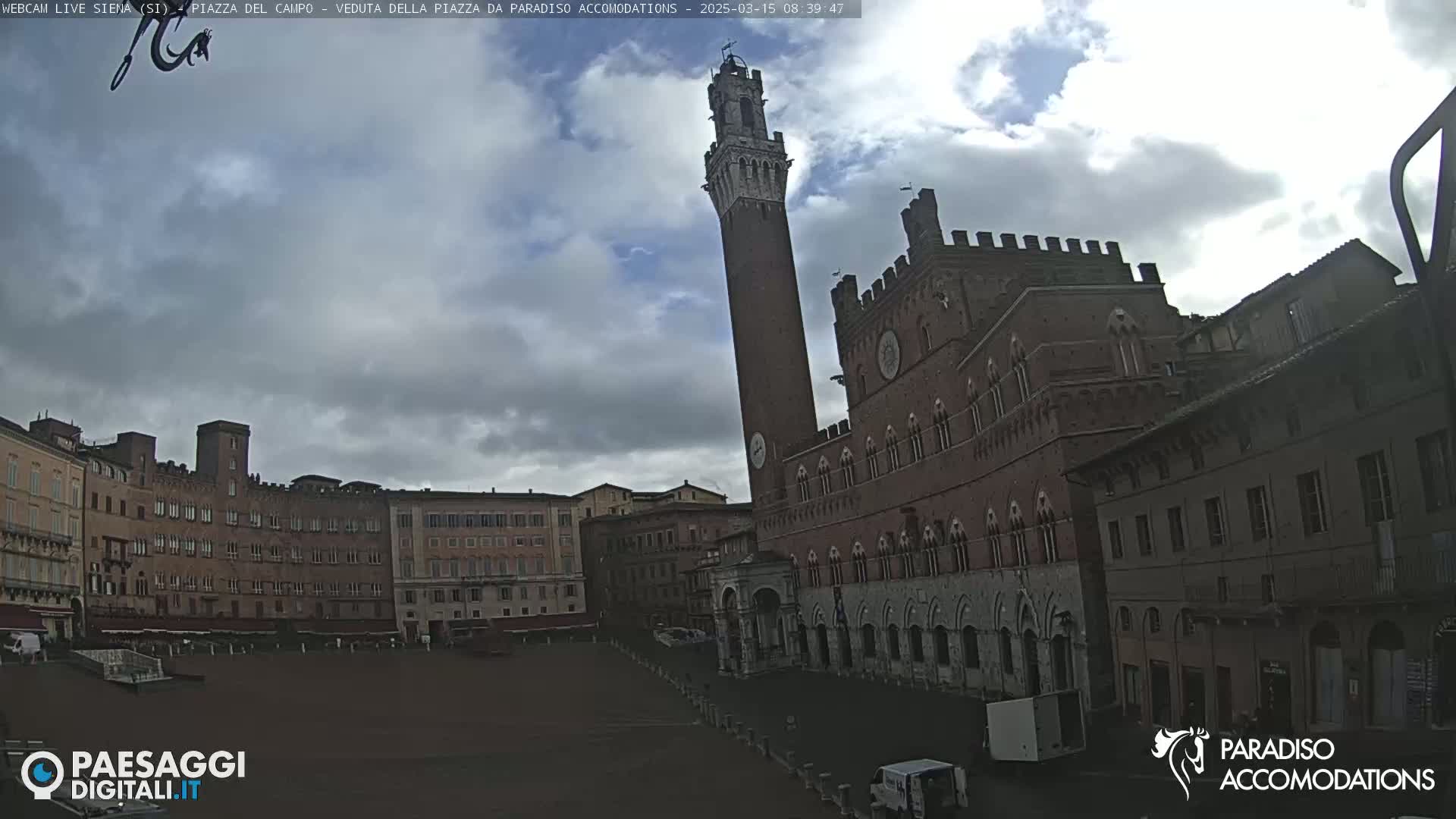 A large, brick public square is shown under a cloudy sky, with a tall, slender tower and a large, ornate brick building prominently featured.