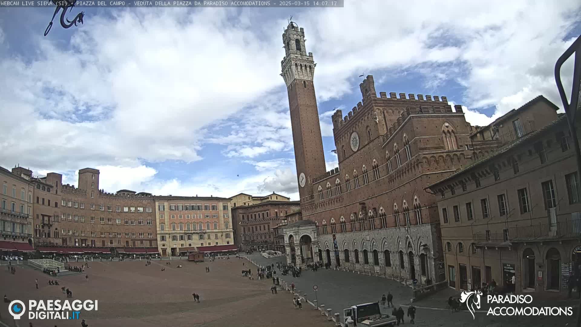 A large, brick piazza in Italy is partially cloudy, with a tall tower and palace visible.
