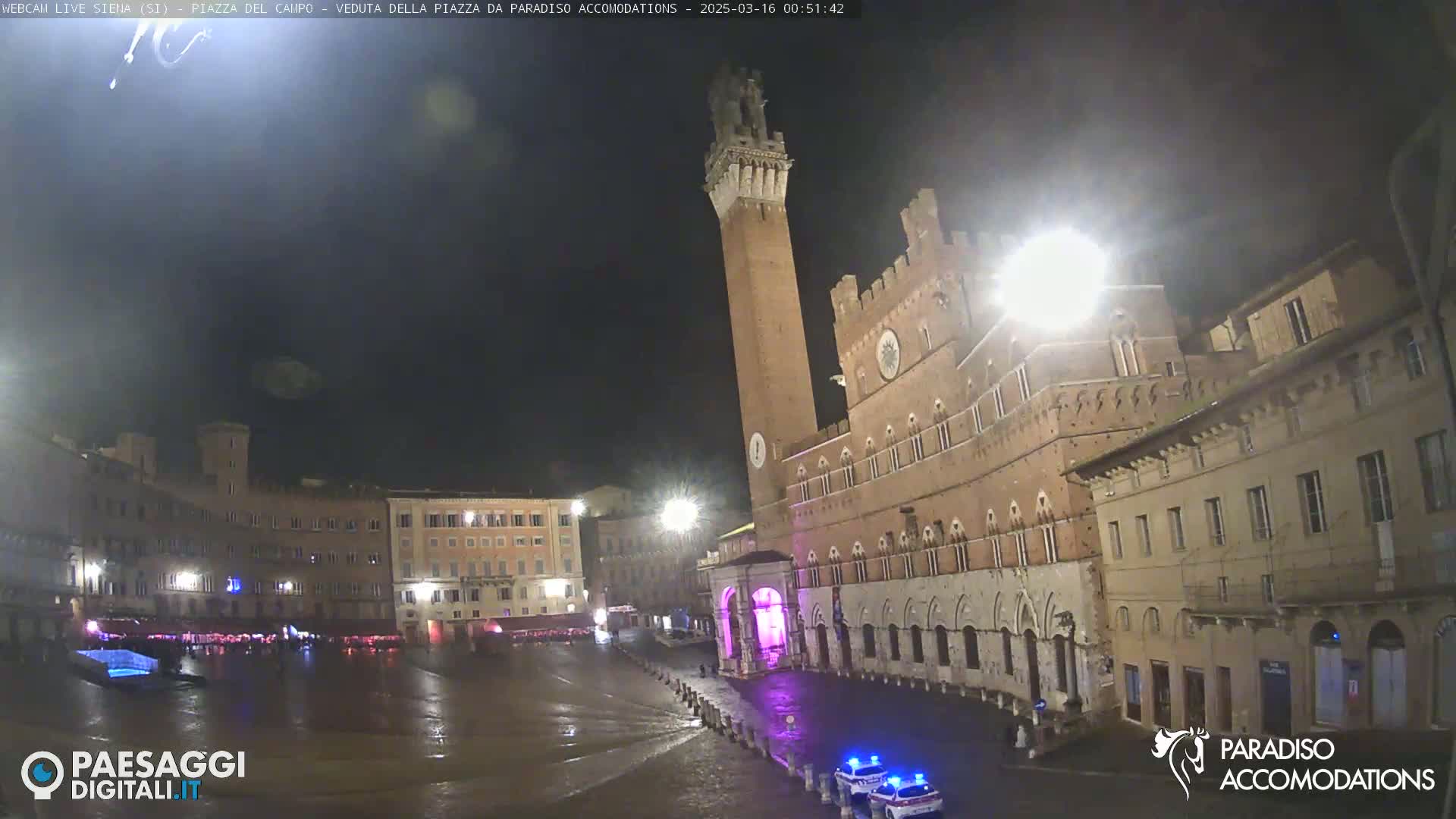 A nighttime view of a large, brick piazza in Italy, illuminated by streetlights, with a tall tower and buildings surrounding a wet, reflective square.