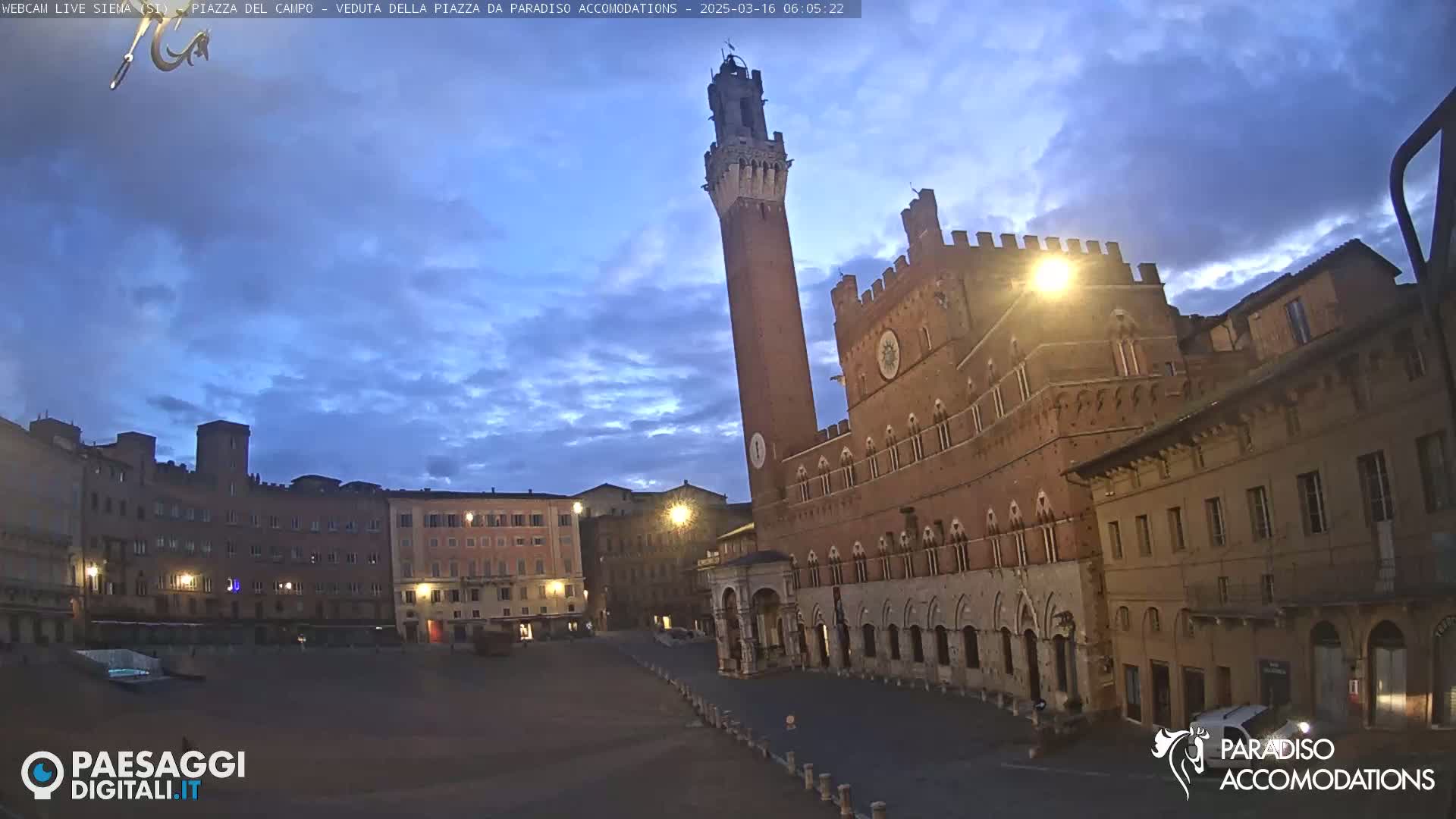 A large, empty plaza in a European city at twilight, featuring a tall brick tower and adjacent medieval-style buildings under a mostly clear sky.