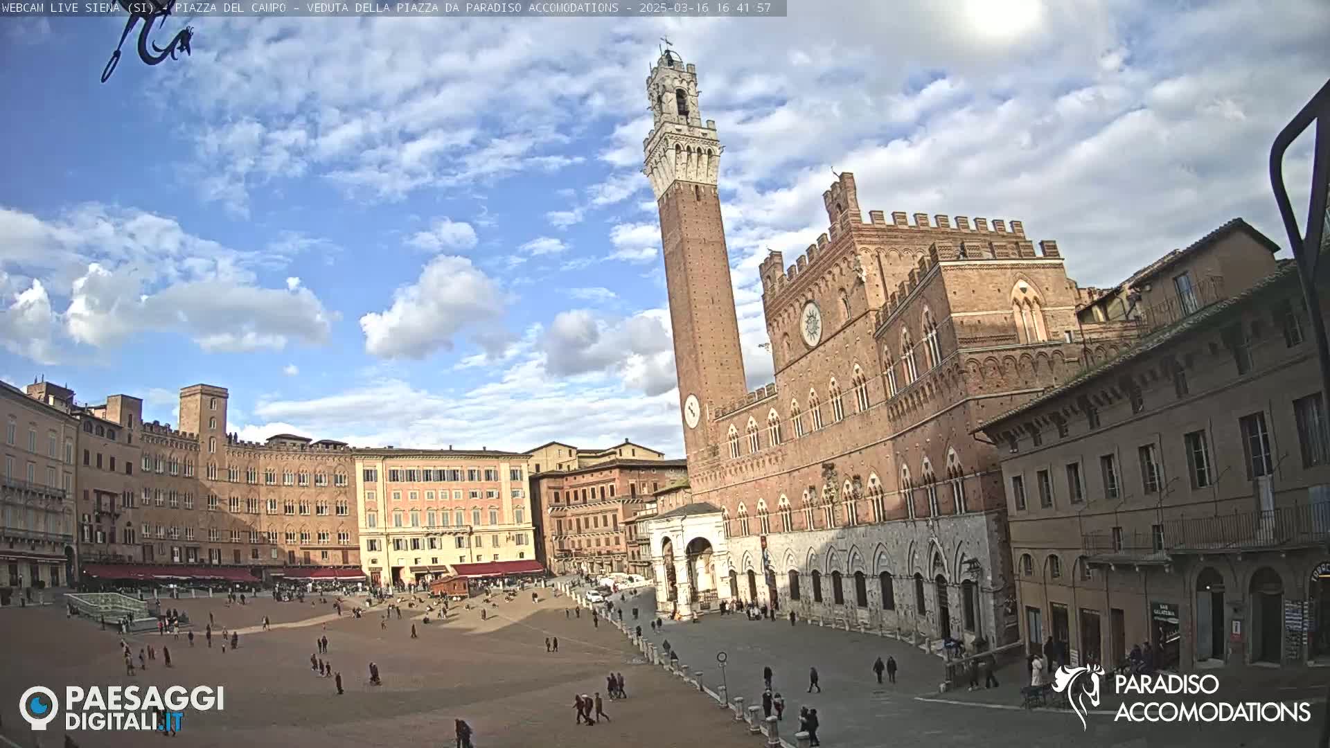 A large, sunny town square is surrounded by medieval-style brick buildings, one prominently featuring a tall tower.