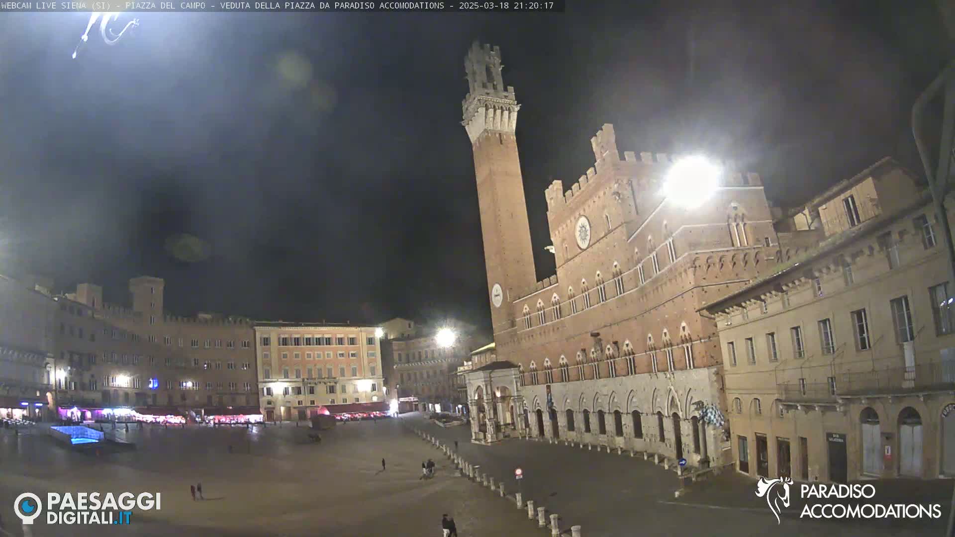 A nighttime view of a large, mostly empty plaza in a European city, featuring a tall brick tower and surrounding historical buildings under a dark sky.