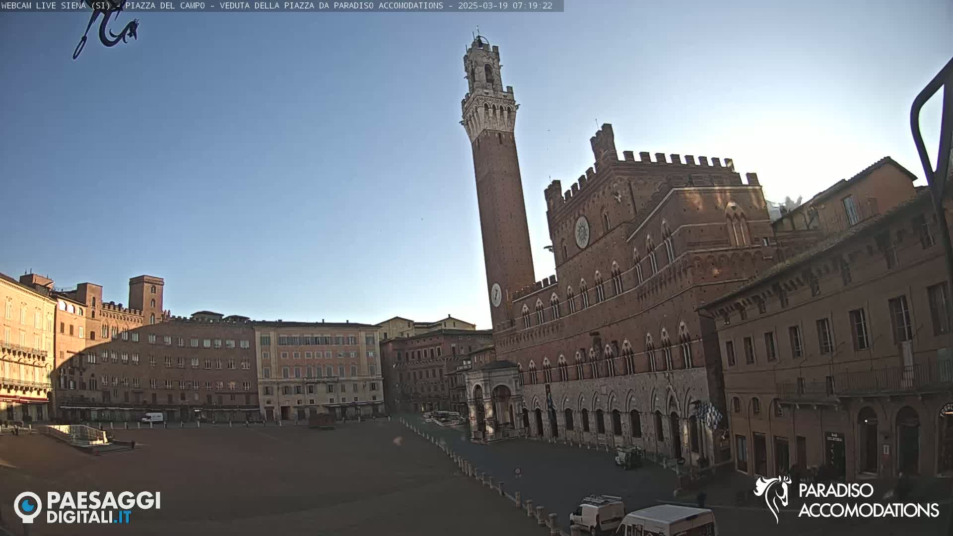 A large, mostly empty, brick plaza is situated in front of a tall, slender brick tower and a large, ornate brick building under a clear, bright sky.