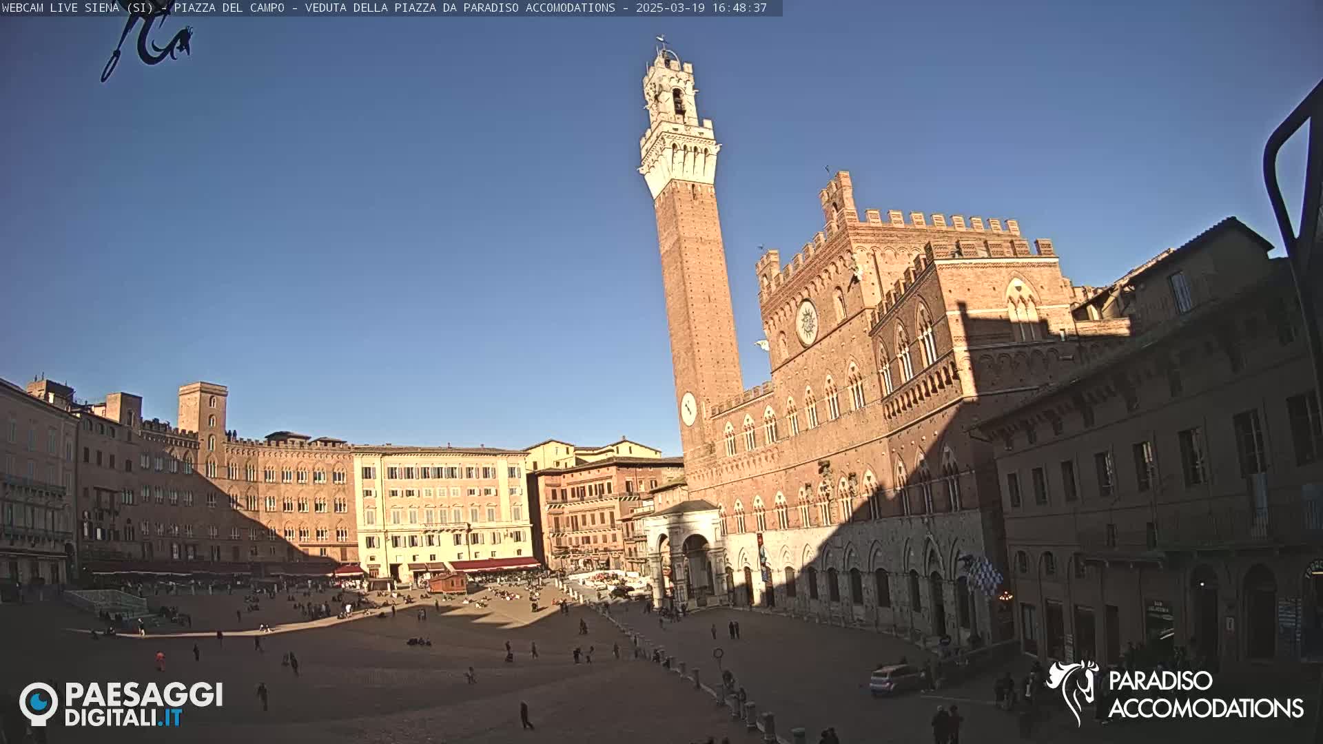 A large, mostly empty, brick plaza is surrounded by medieval-style buildings under a clear blue sky.
