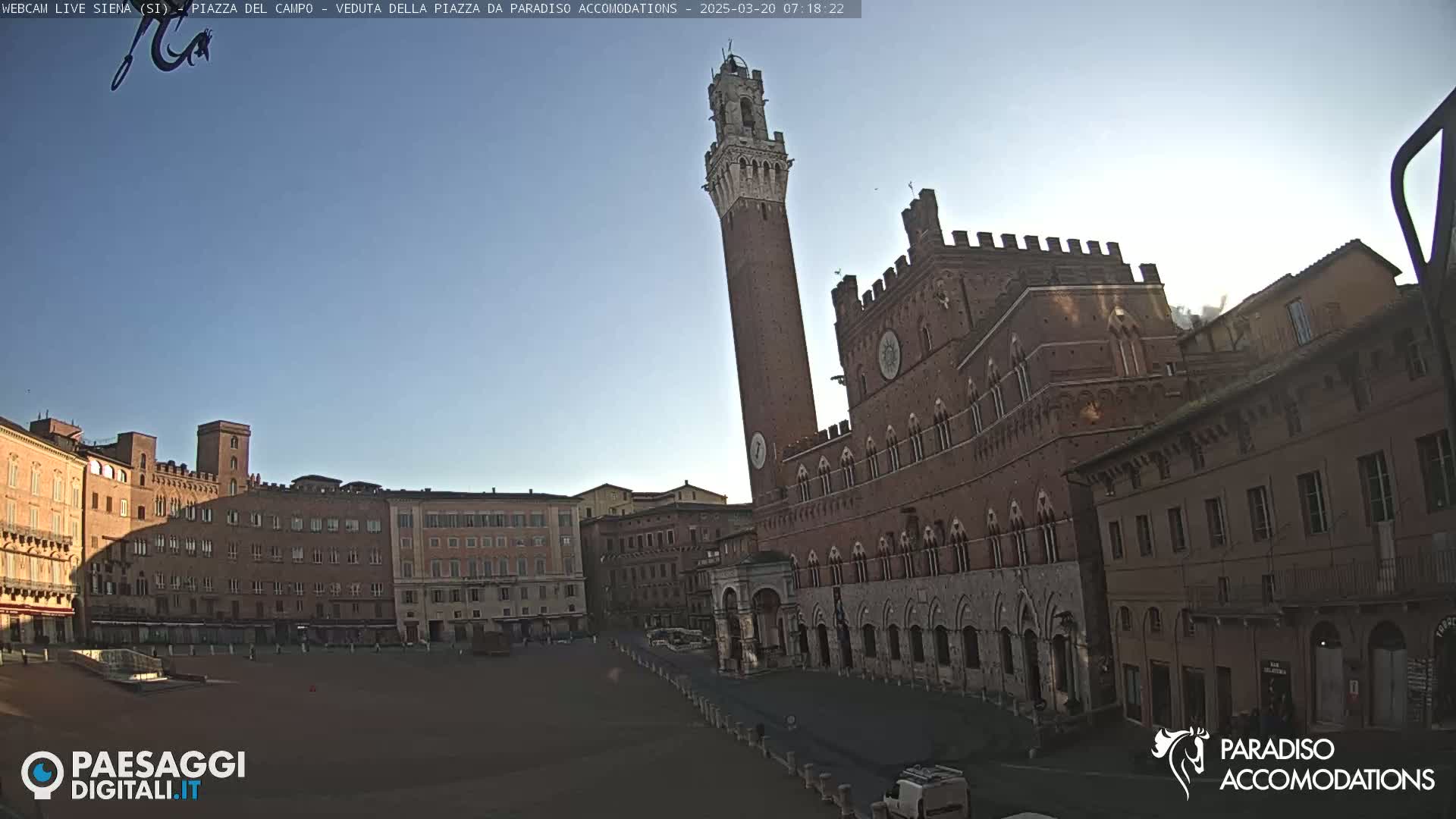 A large, empty plaza in front of a tall brick tower and a long, ornate brick building under a clear, bright sky.
