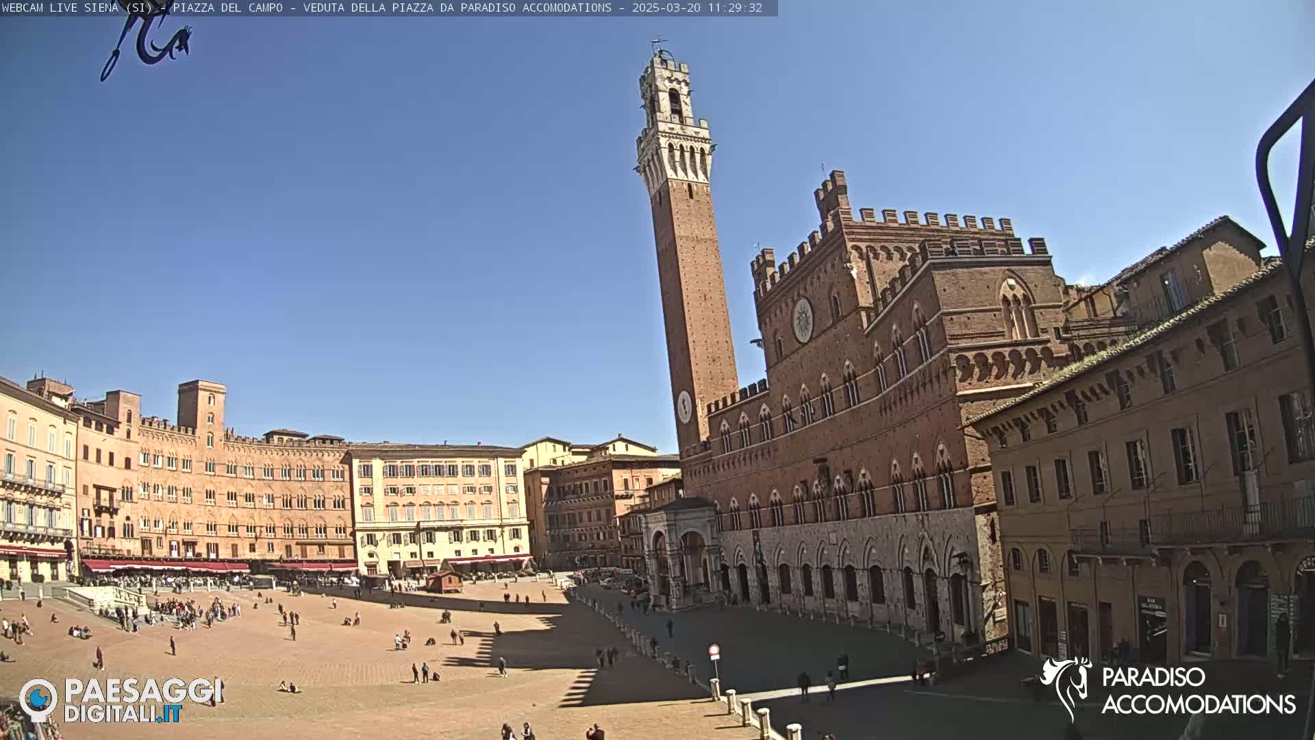 A large, brick plaza is shown on a sunny day, surrounded by medieval-style buildings, including a tall tower.