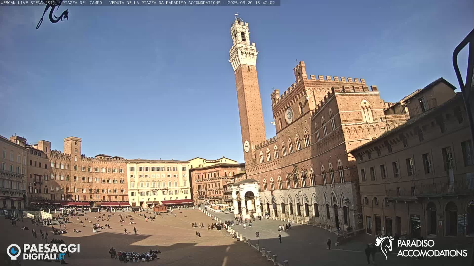 A large, sunlit piazza in Siena, Italy, is surrounded by brick buildings, including a tall tower and a grand palazzo.