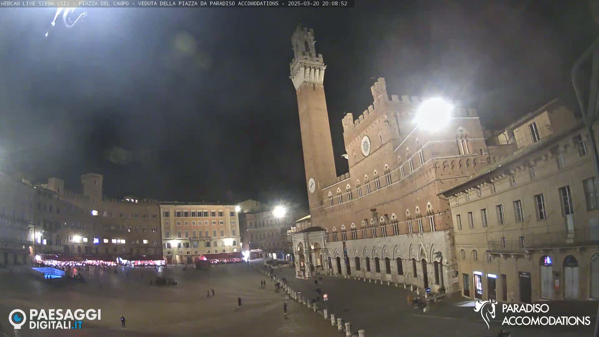 A nighttime view of a large, mostly empty town square surrounded by historic buildings, one of which is a tall brick tower.