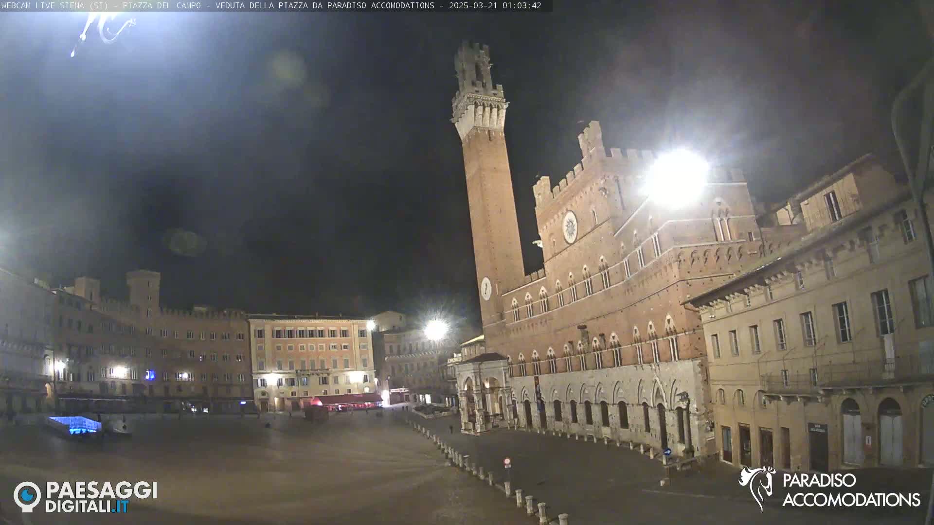 A nighttime view of a large, empty town square in Italy, dominated by a tall brick tower and surrounding medieval-style buildings under a dark, clear sky.