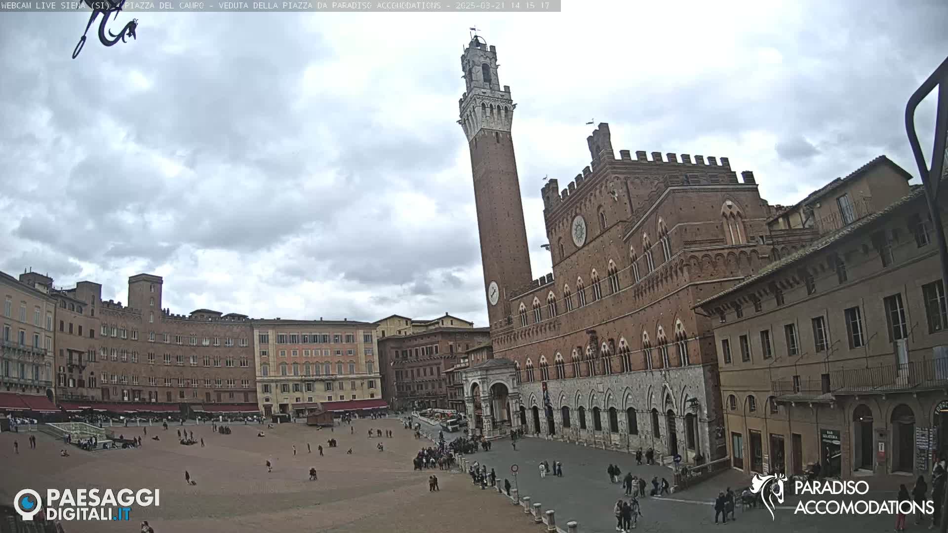 A large, brick plaza is surrounded by historical buildings under a cloudy sky, with a tall, slender tower prominently featured.