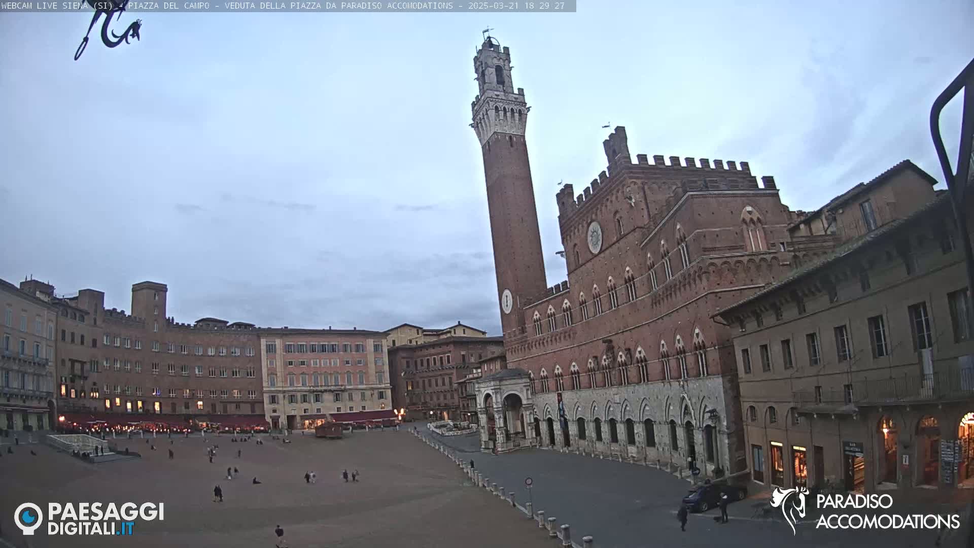 A large, mostly empty plaza in a historic city, under a cloudy, twilight sky, is surrounded by brick buildings, including a tall tower and a grand palace.