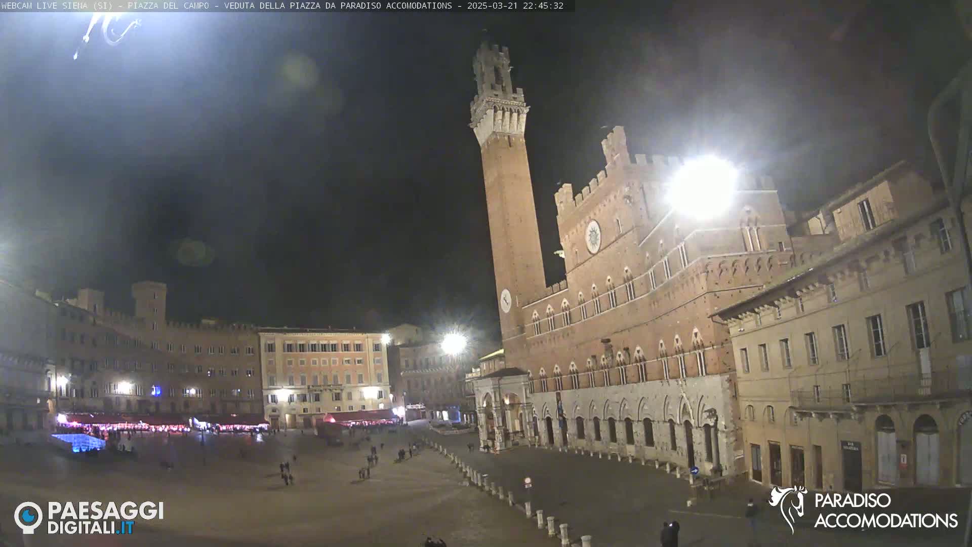 A nighttime view of a large, mostly empty plaza with medieval-style buildings, including a tall brick tower, under a dark sky.