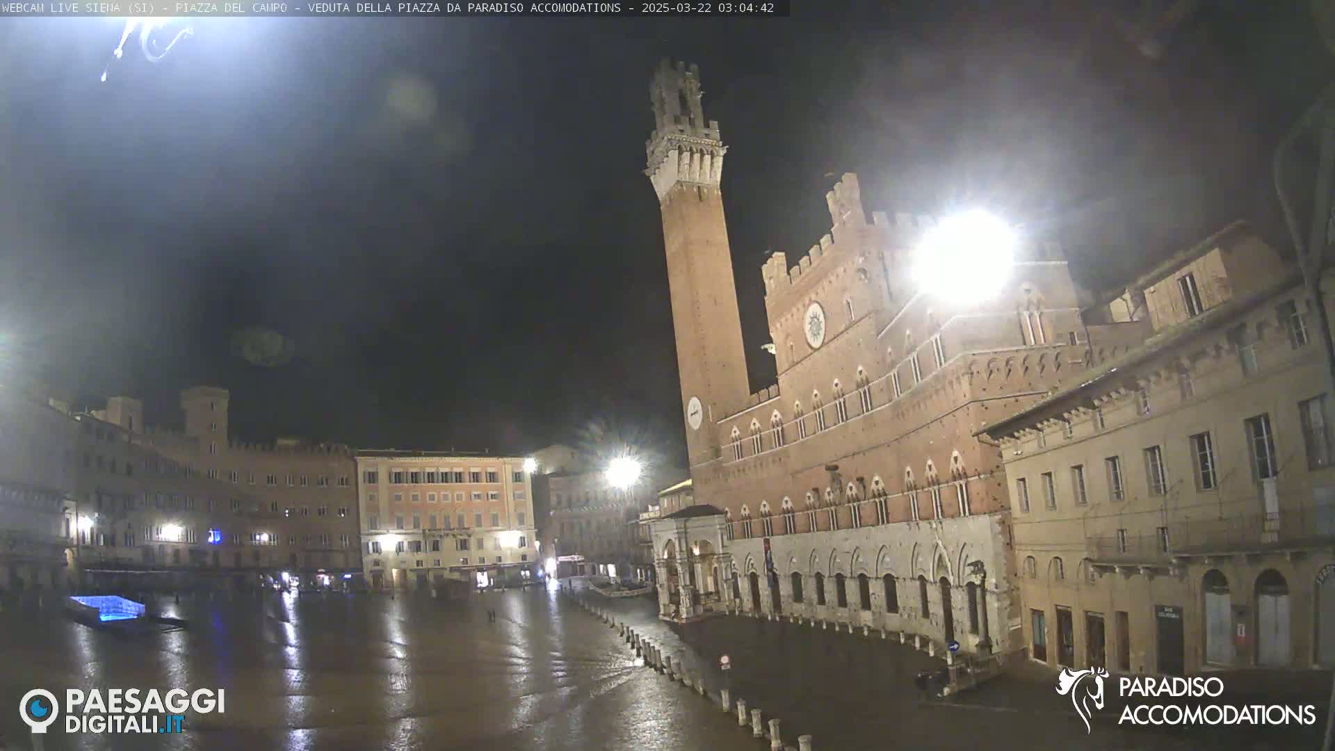 A nighttime view of a large, mostly empty, wet piazza in front of a tall brick tower and adjacent buildings.