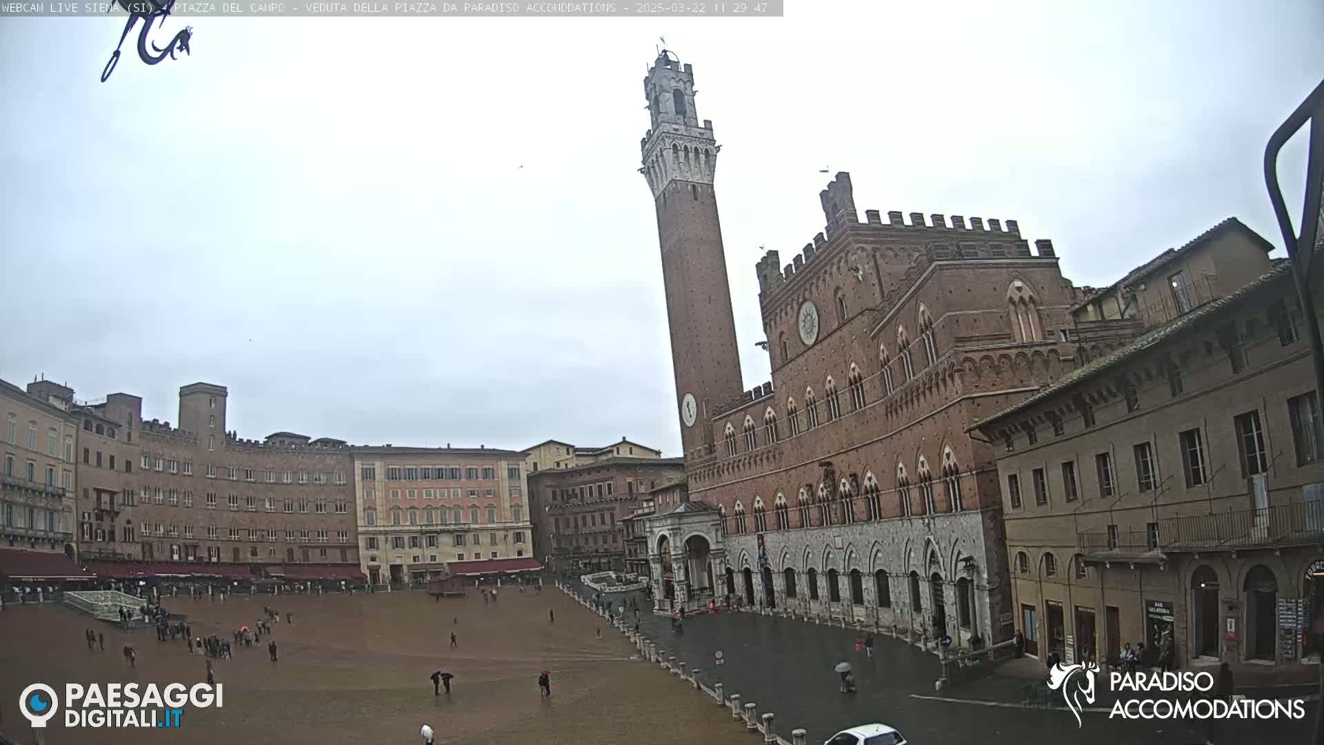A large, brick piazza in a European city is shown on an overcast day, with a tall, slender tower and a grand, ornate building dominating the scene.