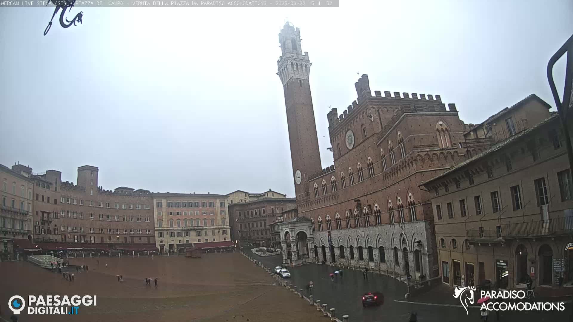 A large, brick piazza in Siena, Italy, is shown on a rainy day, with a tall tower and a grand palace prominently featured.