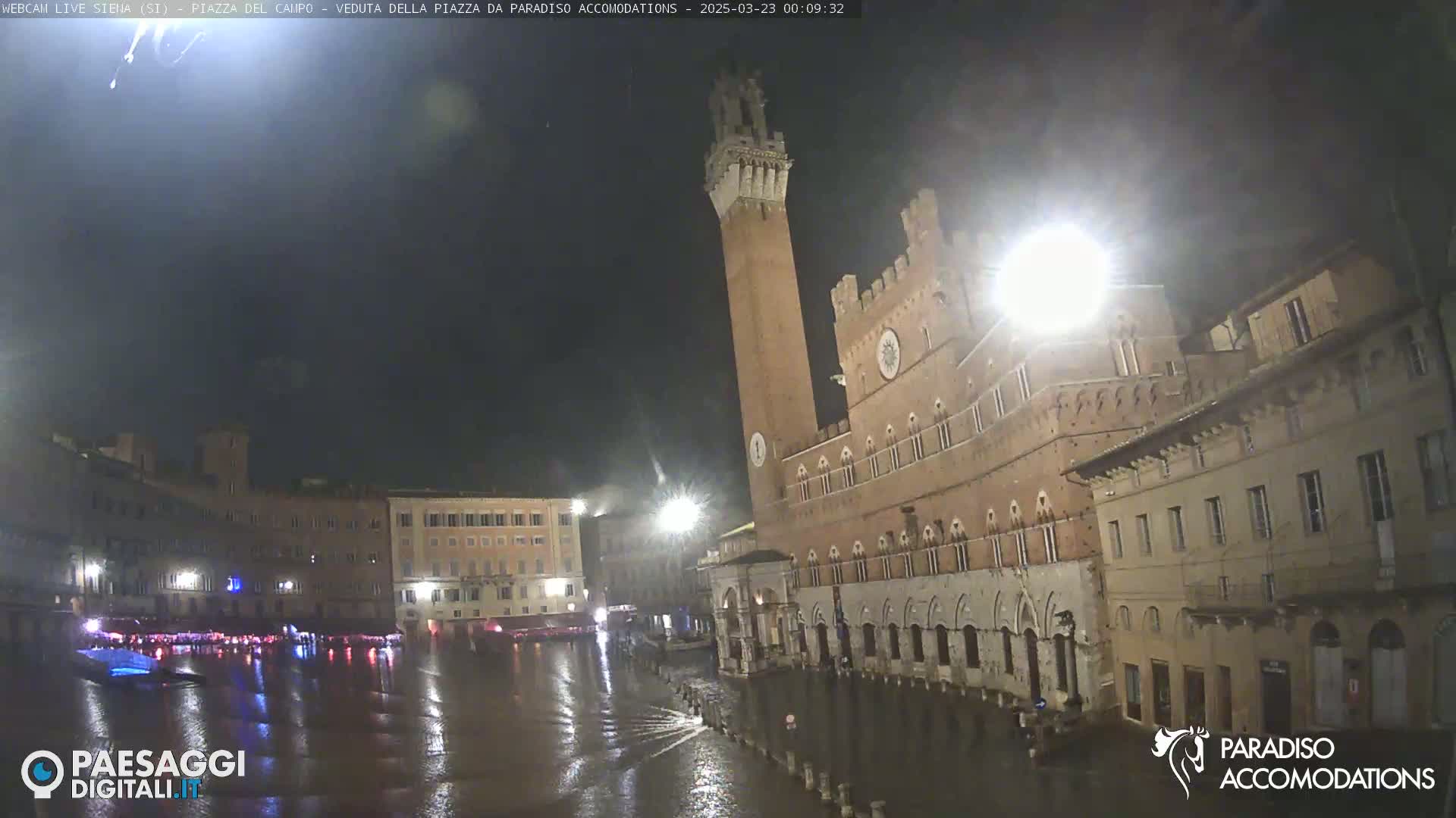 A rain-soaked piazza at night, illuminated by streetlights, features a large brick building with a tall tower, and several other buildings surrounding a reflective, flooded square.
