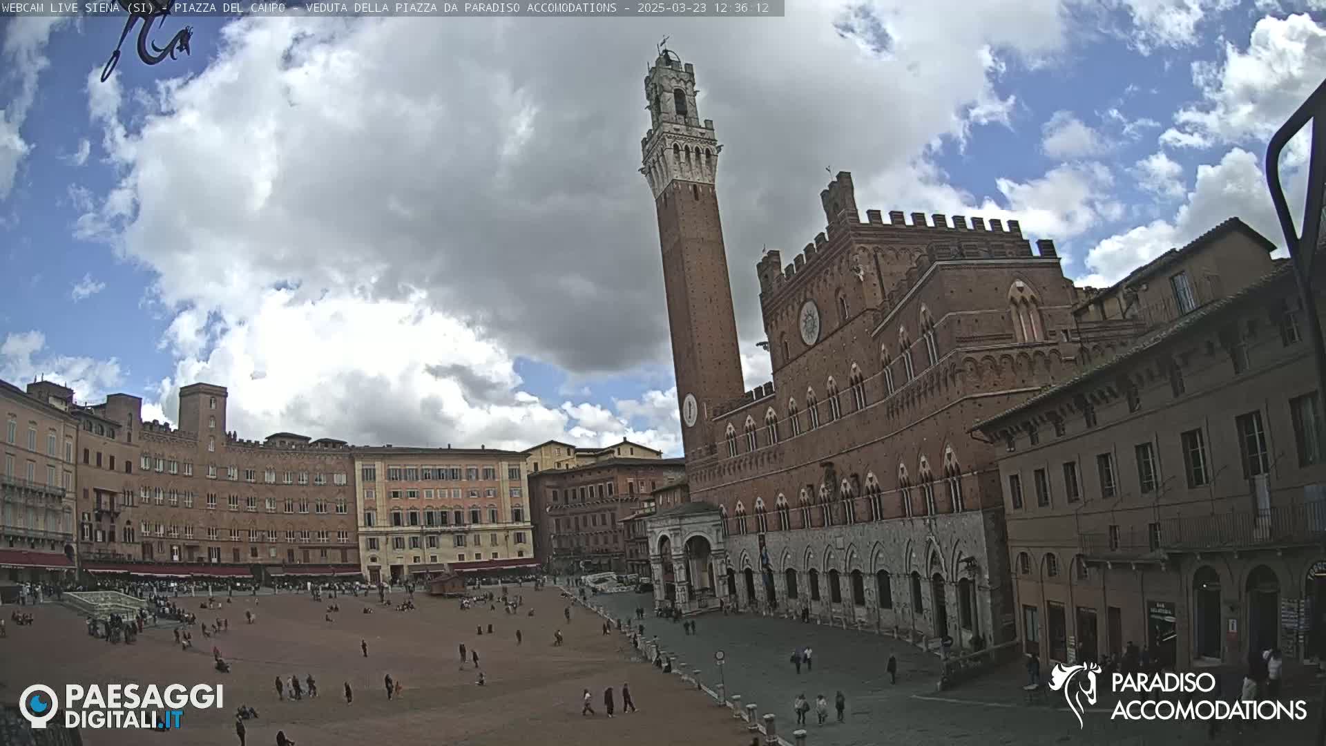 A partly cloudy day in a large, brick-paved plaza surrounded by historic Italian buildings, including a tall tower and a grand palazzo.