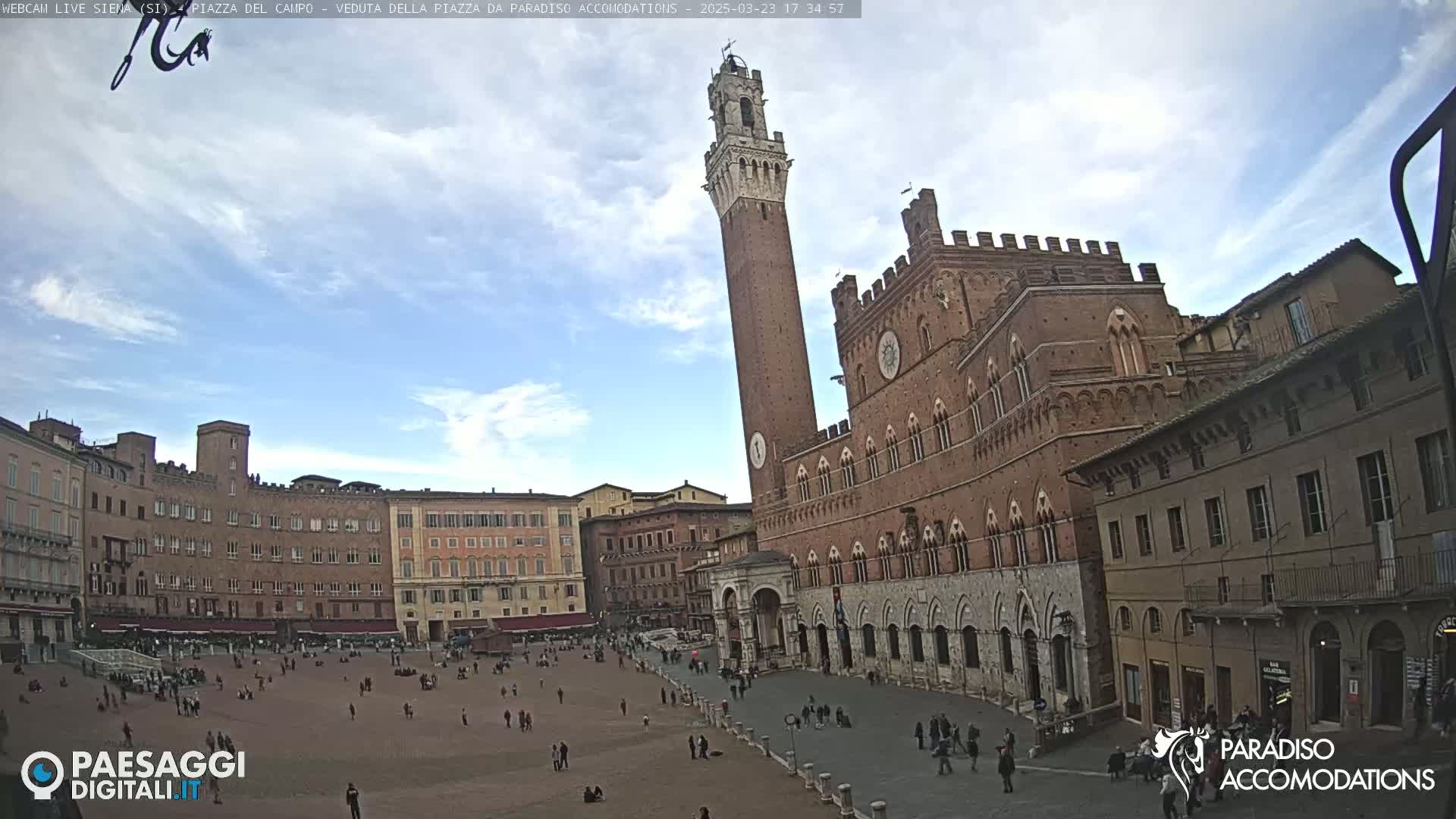 A large, brick public square is populated by a few people, with medieval buildings and a tall tower visible under a partly cloudy sky.