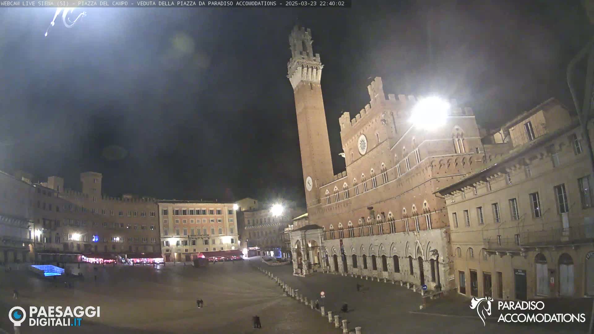 A nighttime view of a large, mostly empty town square surrounded by historic buildings, one prominently featuring a tall tower.