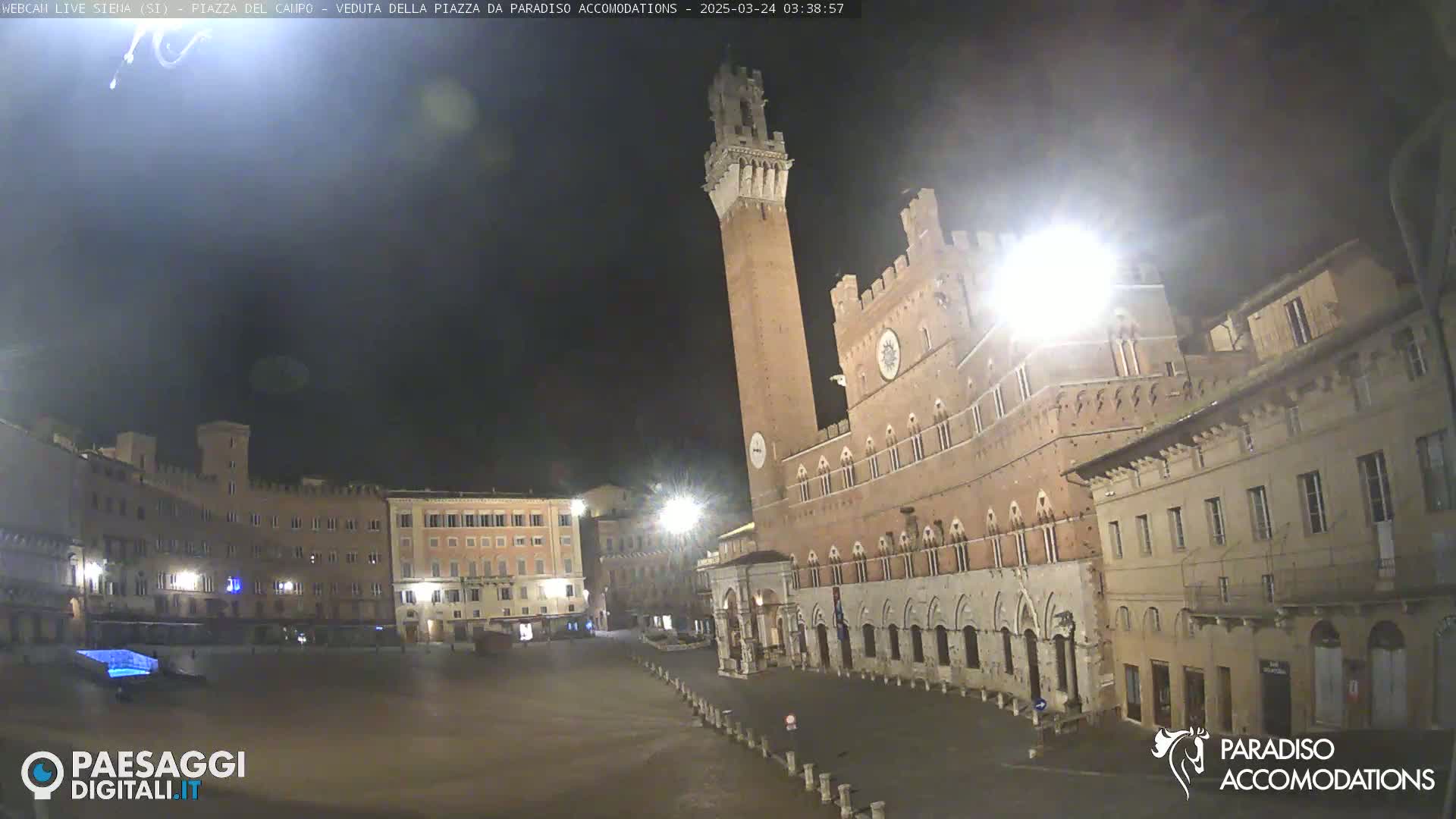 A nighttime view of a large, mostly empty town square in Italy, dominated by a tall brick tower and adjacent historical buildings under a dark sky.