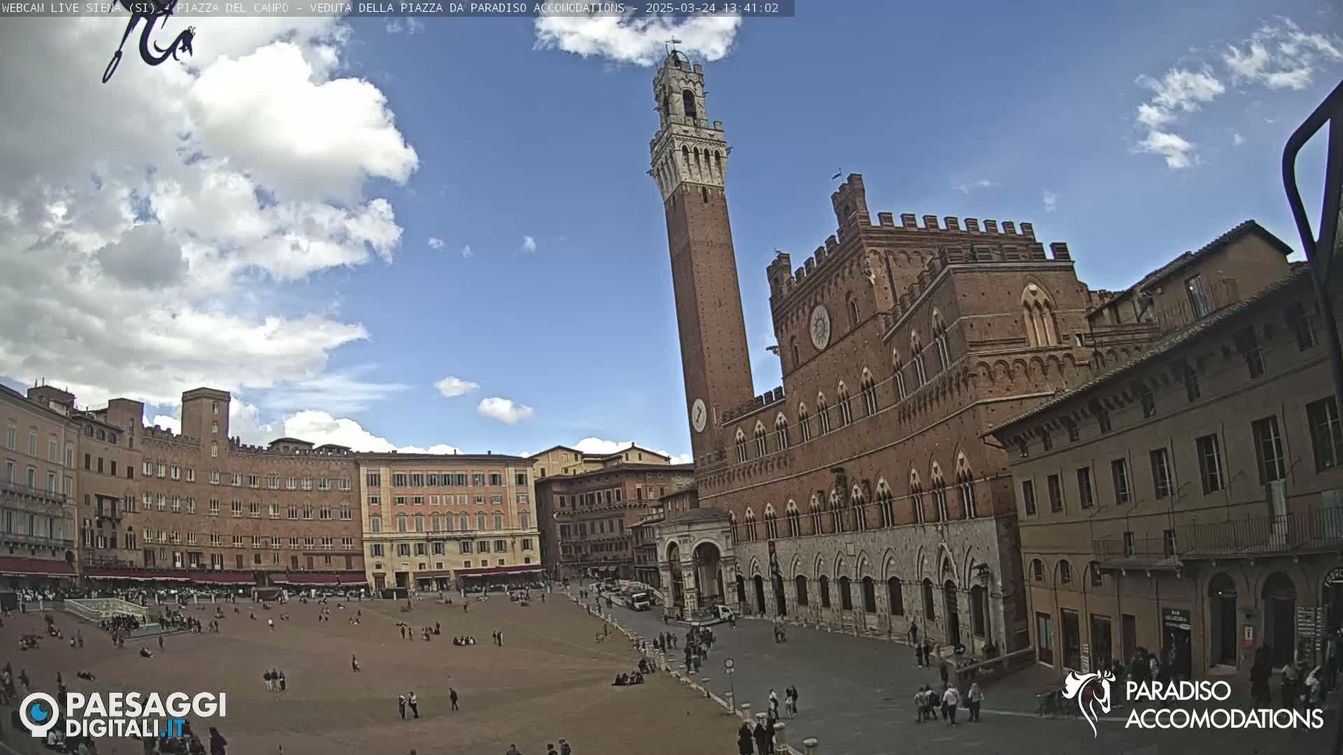 A large, mostly empty plaza in a city is surrounded by brick buildings, including a tall tower and a large palace, under a partly cloudy sky.