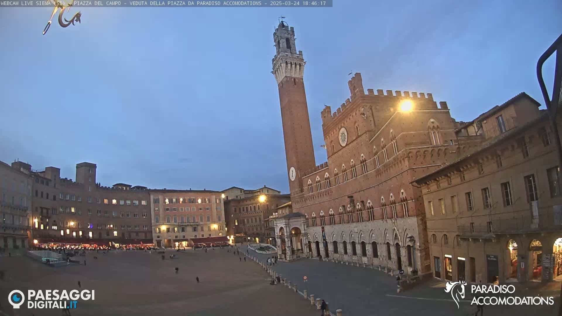 A large, mostly empty, town square at twilight is surrounded by medieval-style buildings, including a tall brick tower.