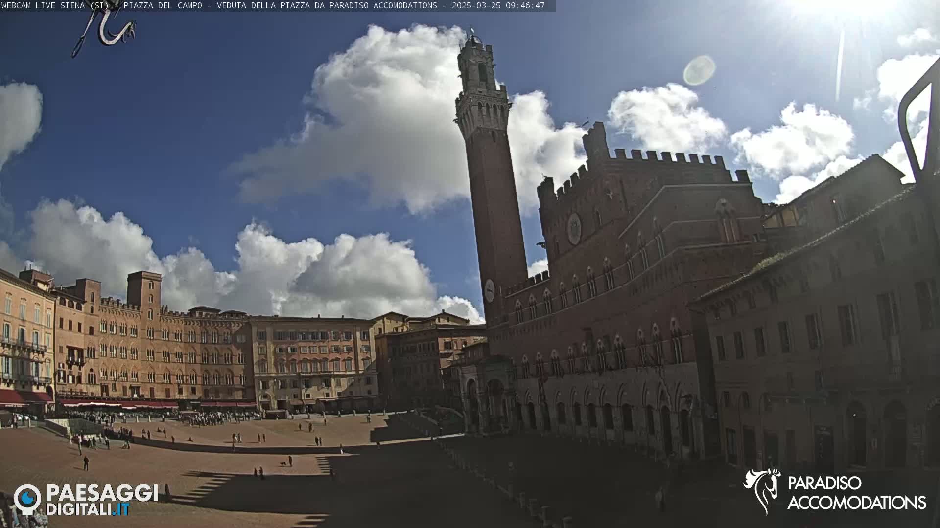 A partly sunny day reveals a large, open plaza surrounded by historic brick buildings, one prominently featuring a tall tower.