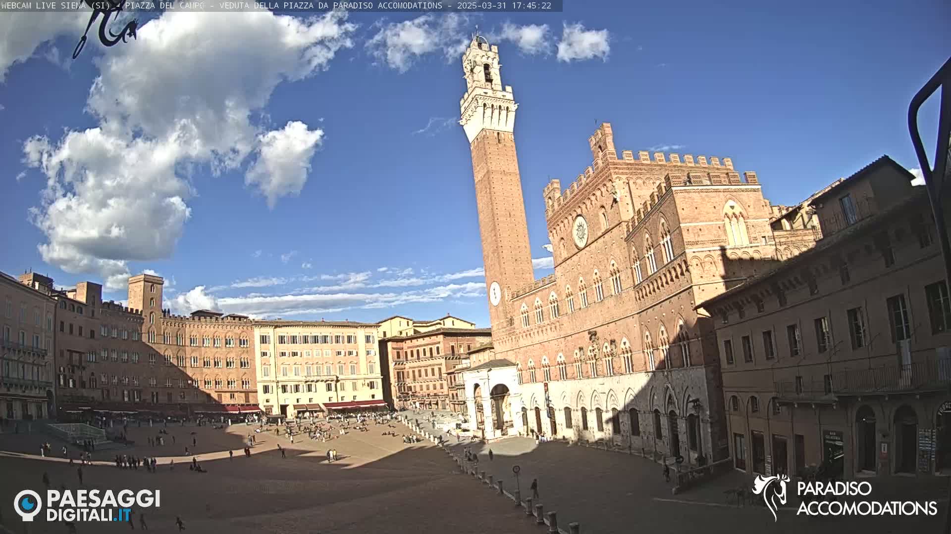 A sunny day in a large, open plaza surrounded by historic brick buildings, including a tall tower and a grand palace, with a few people scattered throughout.