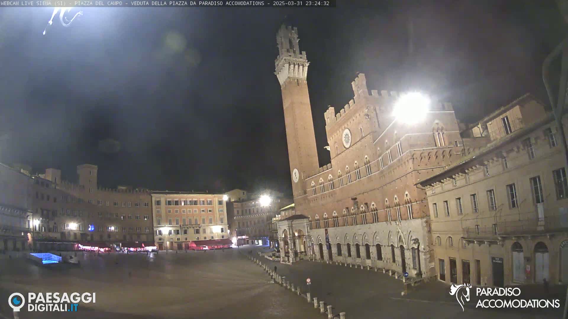 A mostly empty, large, stone plaza at night, featuring a tall brick tower and surrounding medieval-style buildings under a dark sky.
