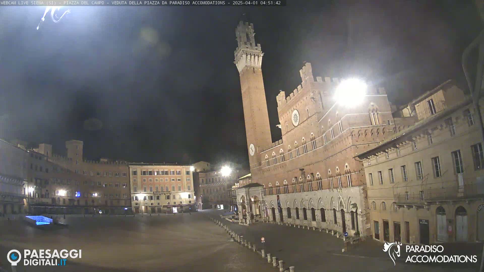 A nighttime, empty, cobblestone town square in Italy, dominated by a tall brick tower and surrounding historic buildings under a dark, clear sky.