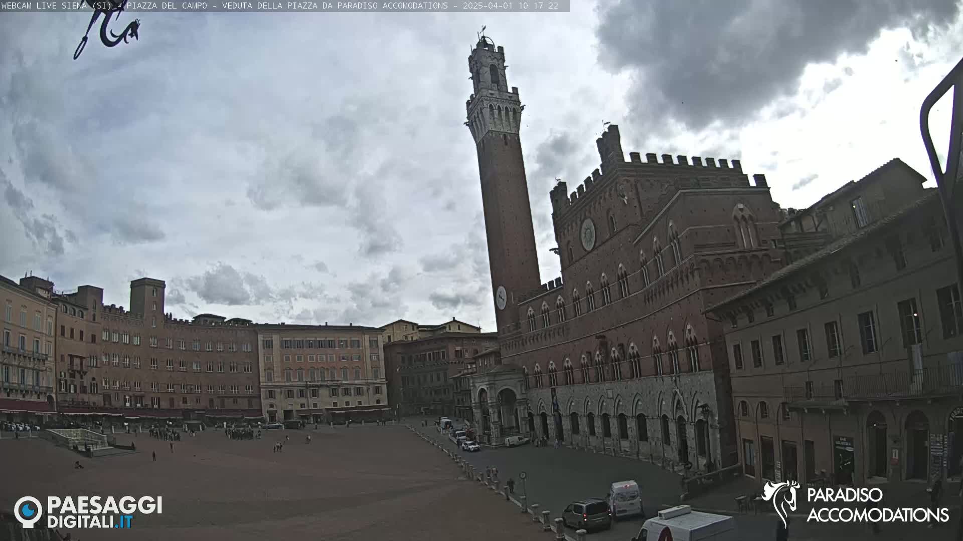 A large, brick public square in a European city, under a mostly cloudy sky, is surrounded by medieval-style buildings, one of which has a tall, slender tower.