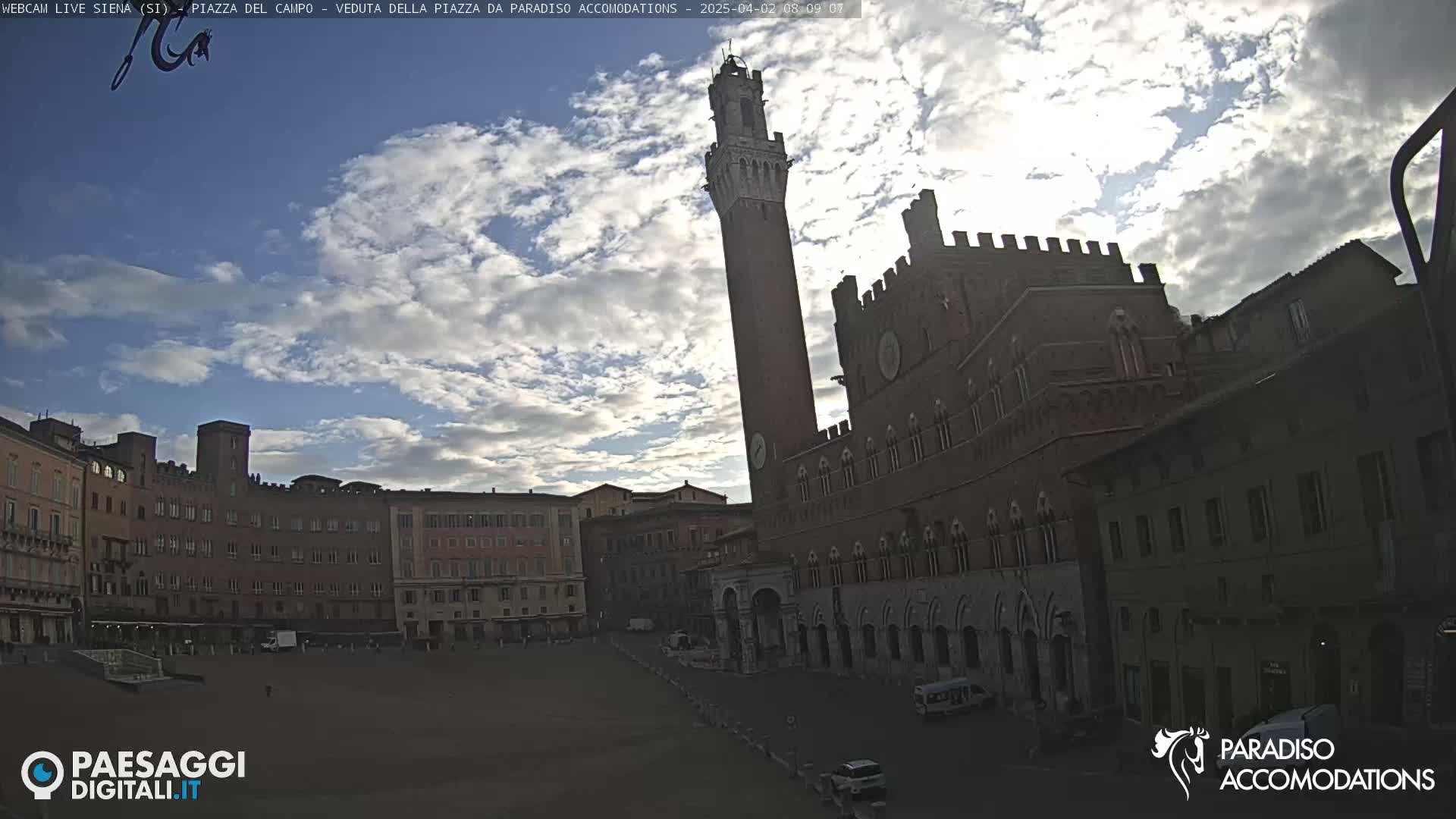 A large, mostly empty plaza is surrounded by historic brick buildings, including a tall tower, under a partly cloudy sky.