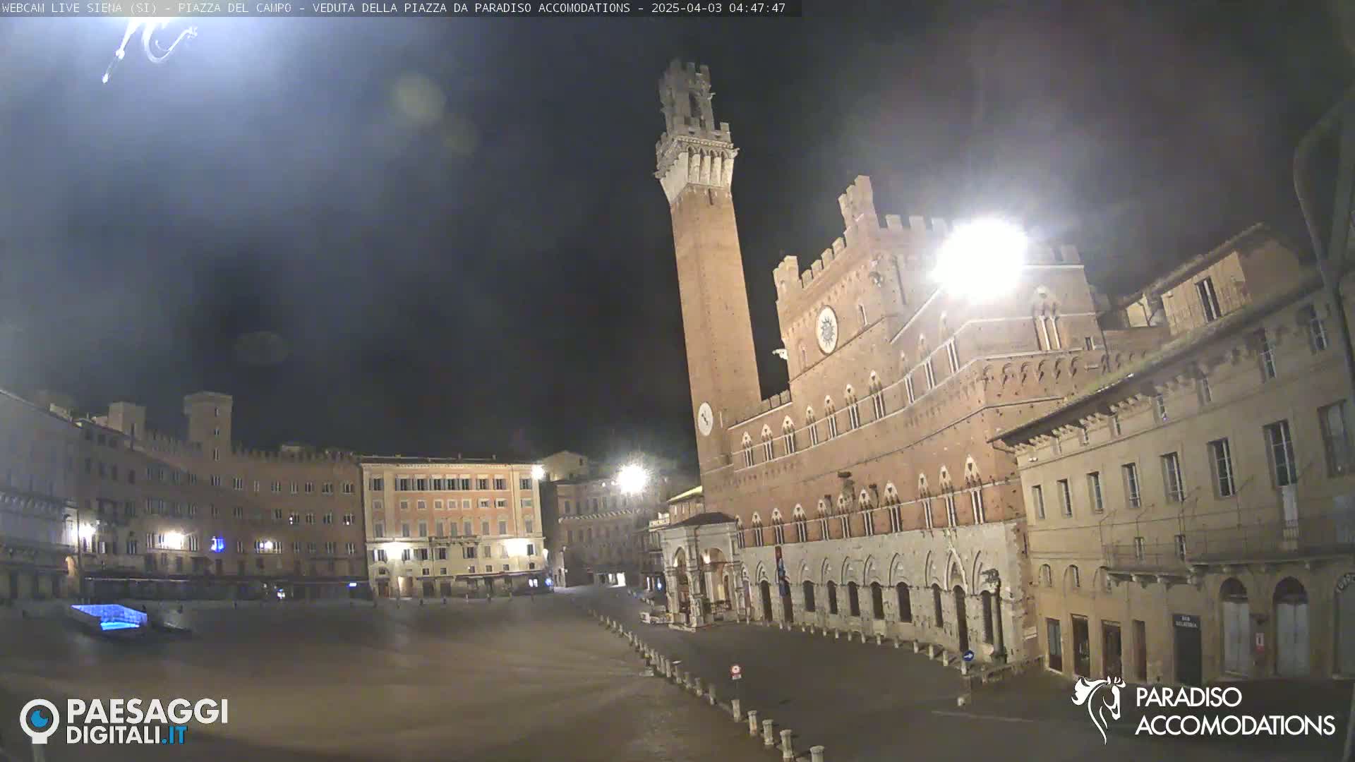 A nighttime view of a large, empty plaza with a tall brick tower and surrounding buildings.