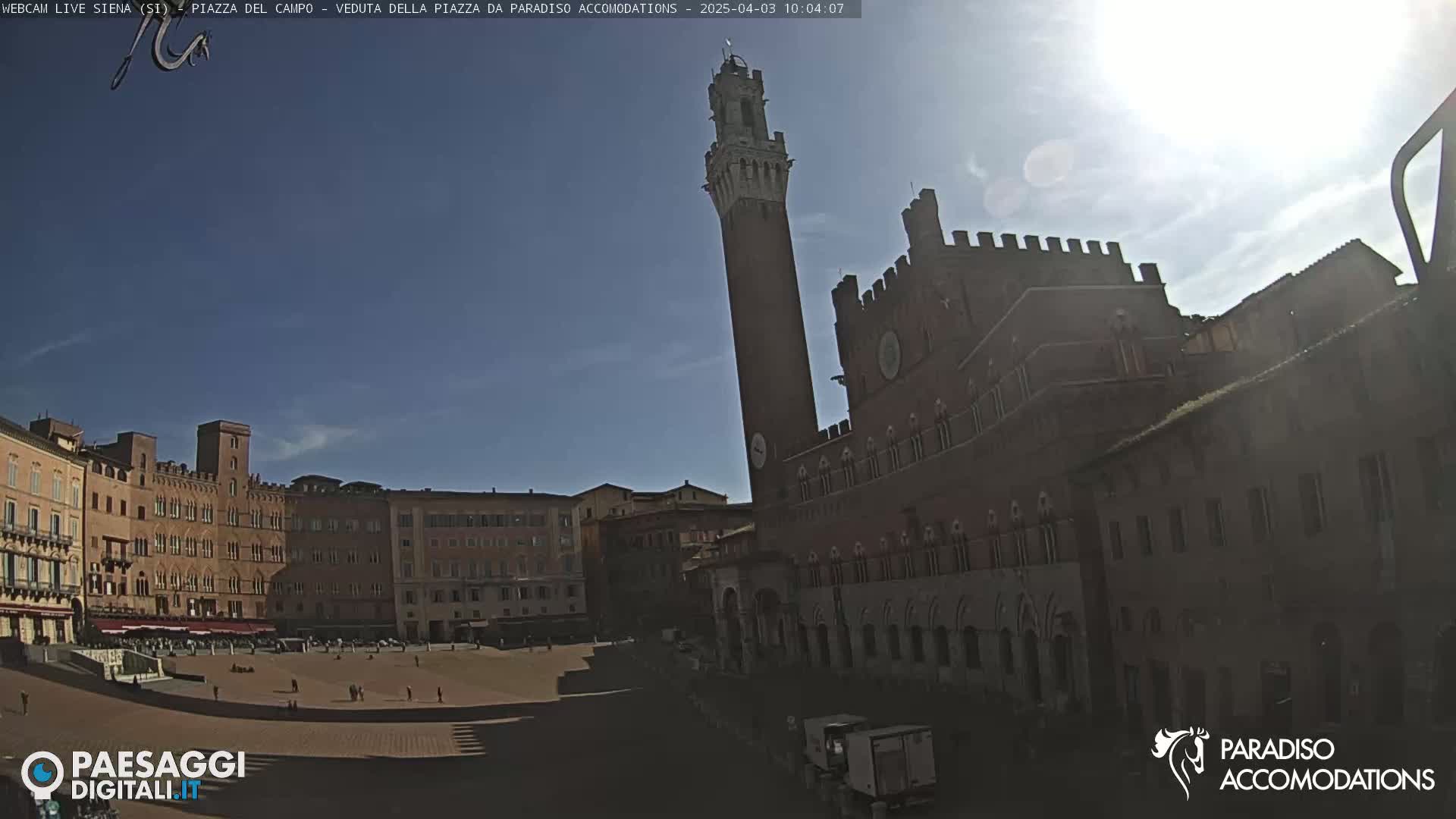 A mostly sunny day reveals a large, brick piazza in Italy, dominated by a tall, slender tower and surrounding medieval-style buildings.