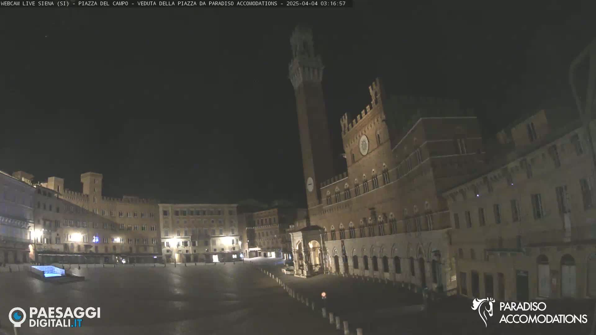 A large, empty town square at night is surrounded by historic, illuminated buildings, one prominently featuring a tall tower.