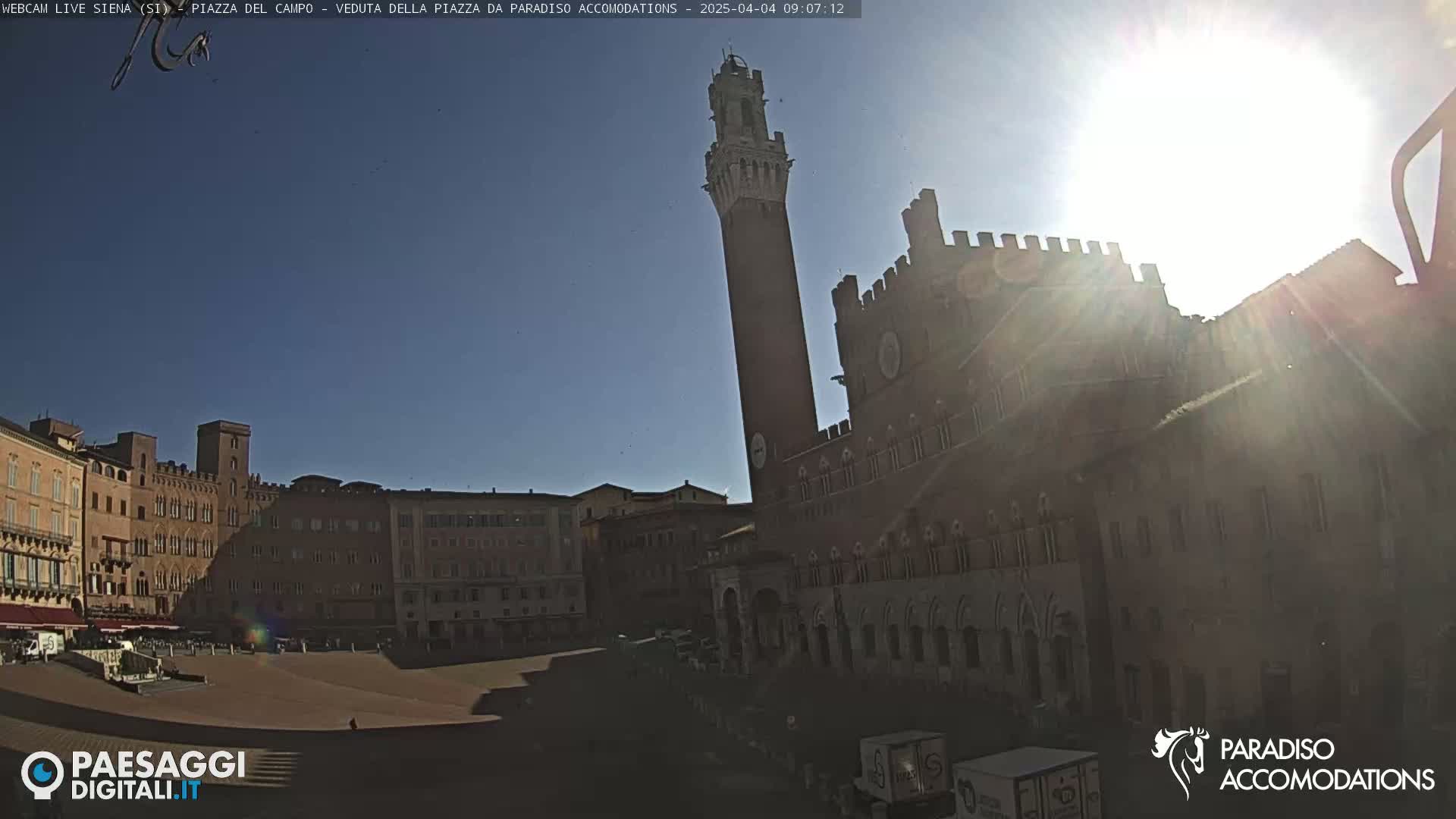 A sunny day reveals a large, mostly empty plaza surrounded by historic brick buildings, including a tall, slender tower.