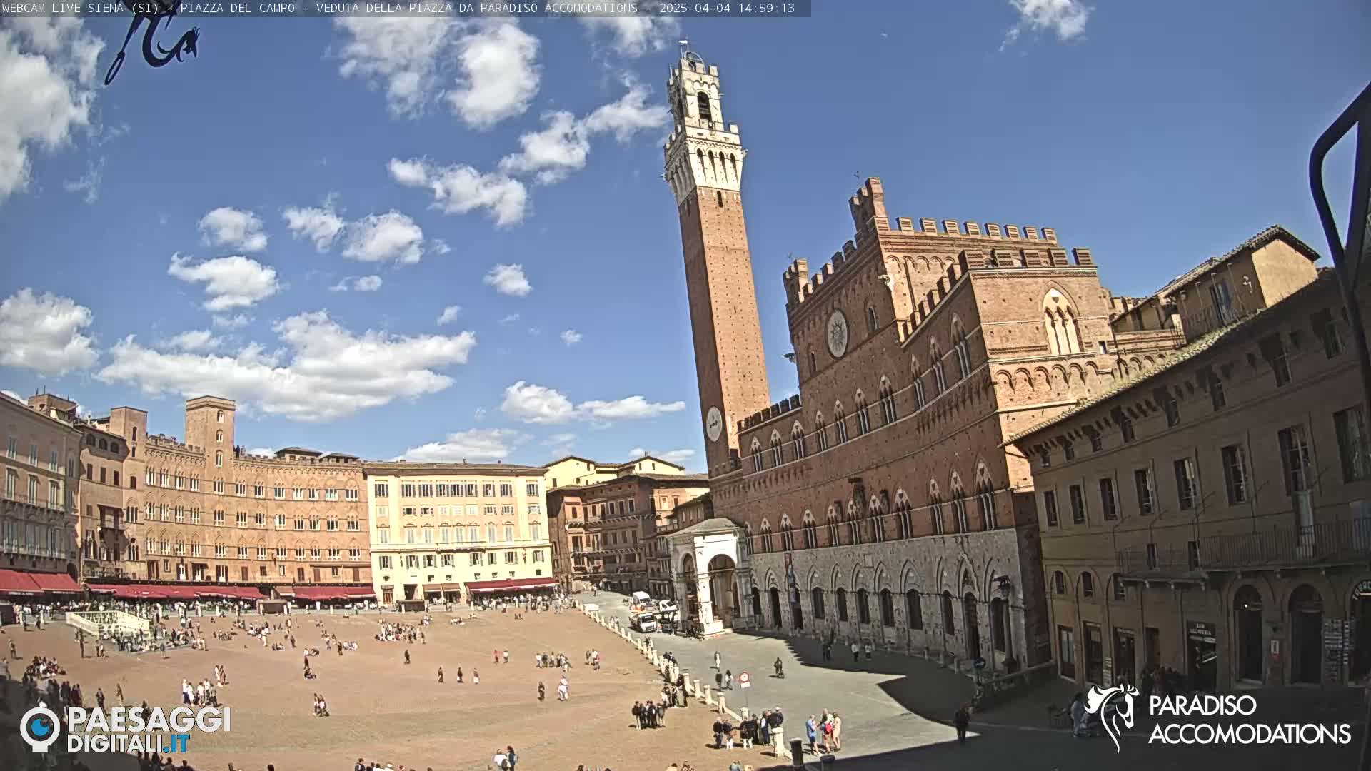 A large, mostly empty plaza is surrounded by brick buildings, including a tall tower, under a partly cloudy blue sky.