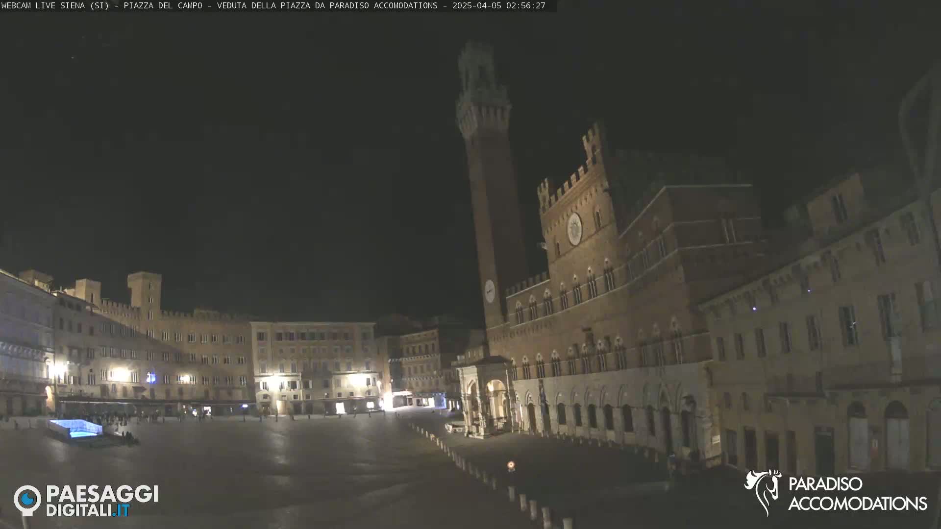 A nighttime view of a large, empty plaza surrounded by illuminated medieval buildings, one featuring a tall tower.