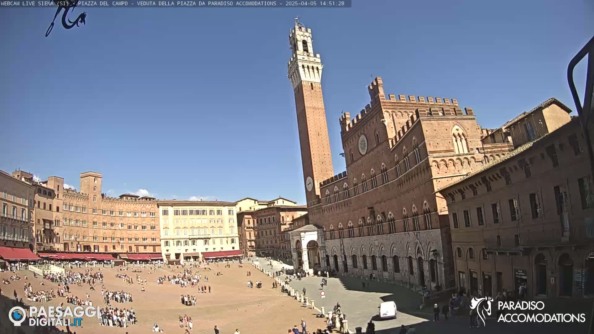 A large, sunlit plaza in Italy is filled with people, surrounded by historic brick buildings, one featuring a tall tower.
