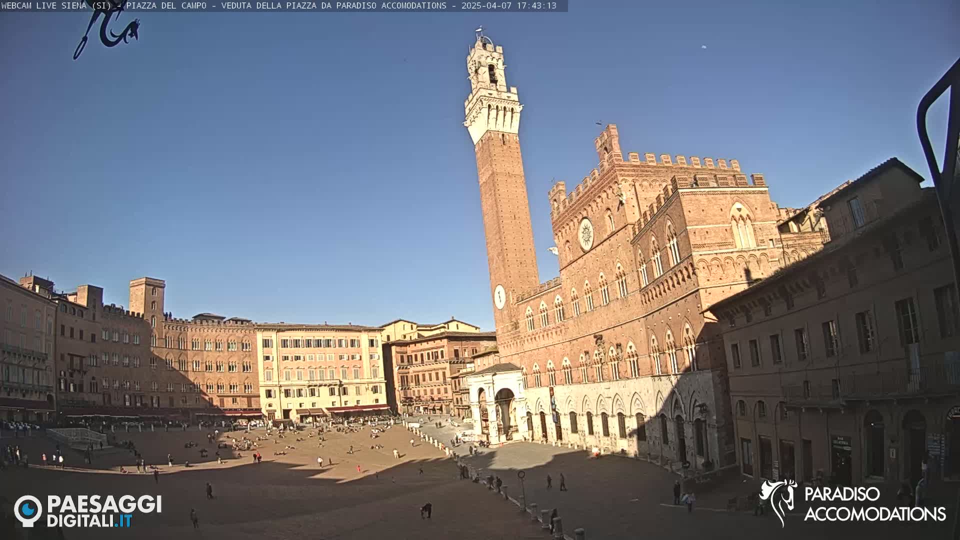 A large, brick piazza with a tall tower and a palace is shown on a sunny day.
