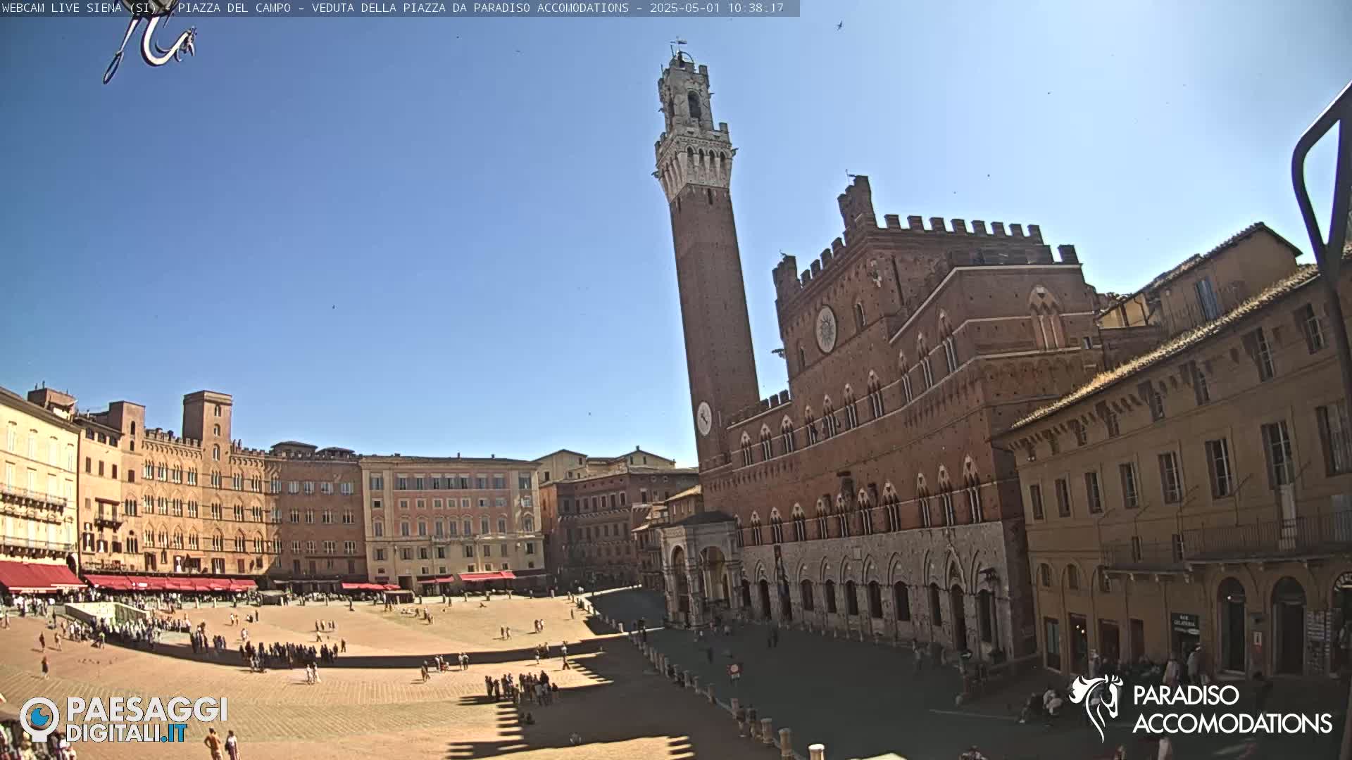A large, brick piazza in bright sunlight, surrounded by medieval-style buildings, including a tall tower.
