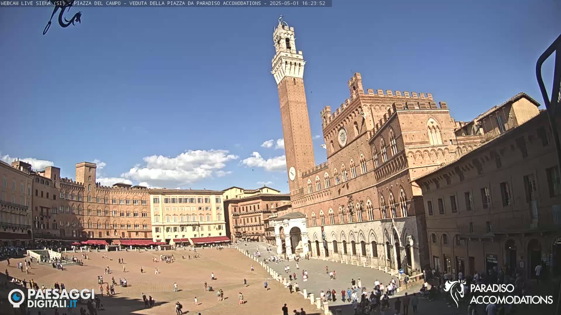 A large, sunny plaza in Italy is surrounded by historic brick buildings, including a tall tower and a grand palace, with people strolling about.