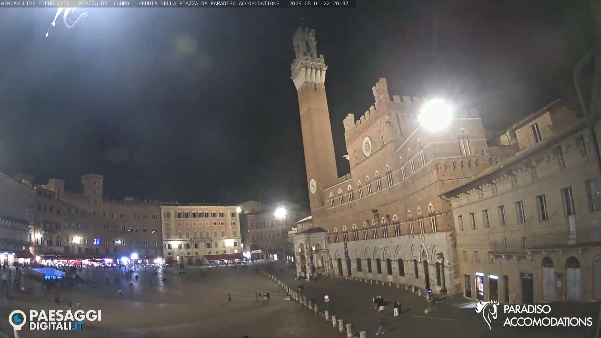 A large, mostly empty town square at night, illuminated by streetlights, is surrounded by historic brick buildings, including a tall, prominent tower.