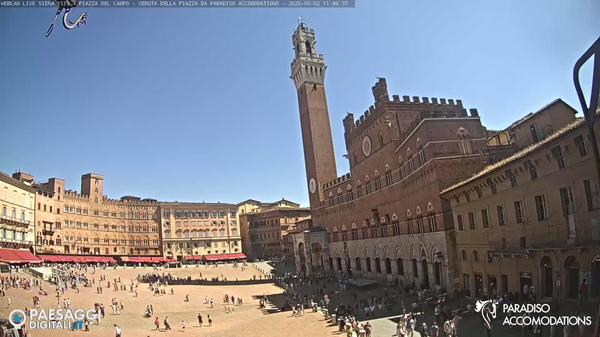A large, mostly empty, square is surrounded by red brick buildings, including a tall tower, under a clear blue sky.