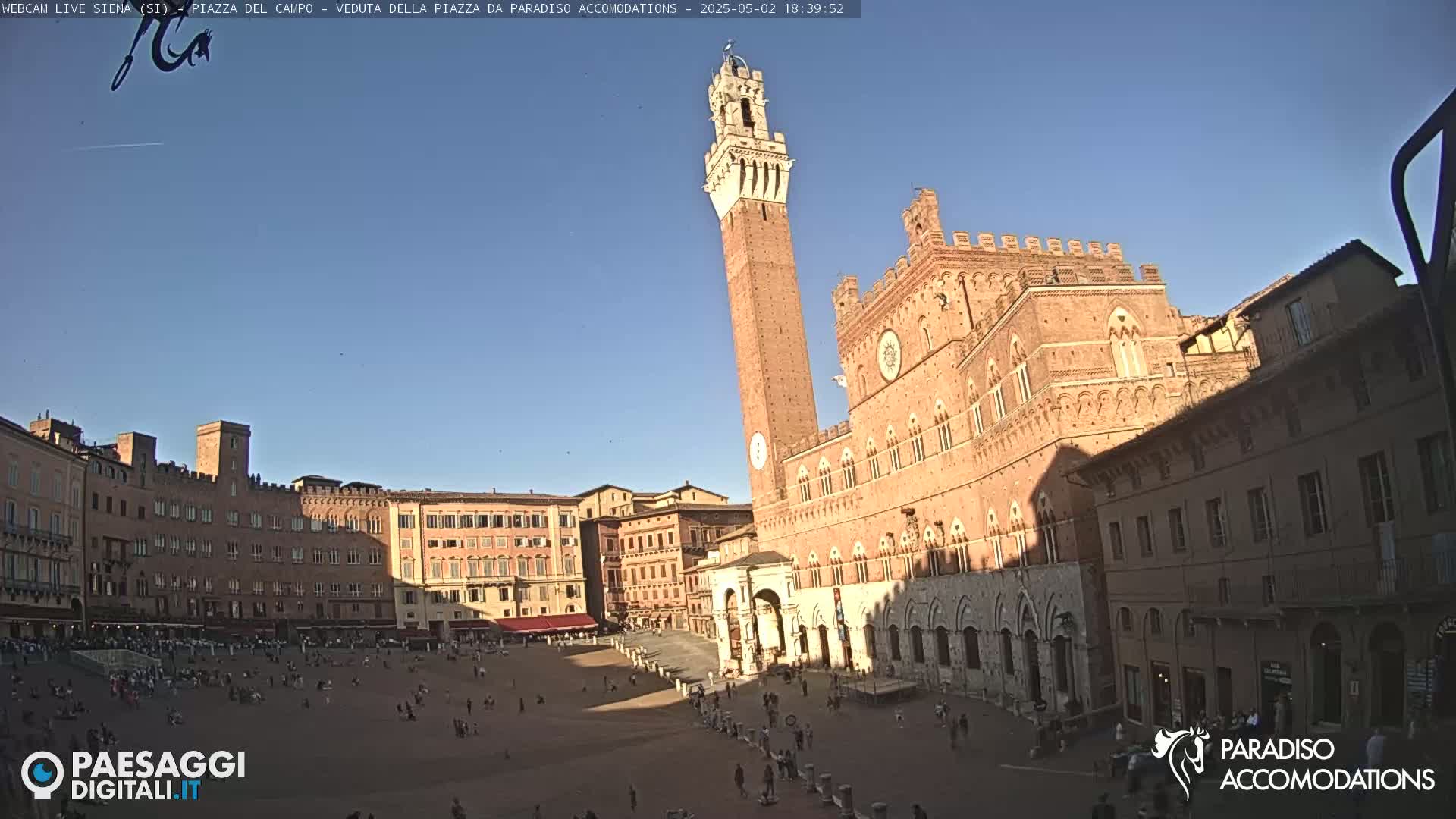 A large, sunlit plaza in a city with several brick buildings, including a tall tower and a large palace, under a clear blue sky.