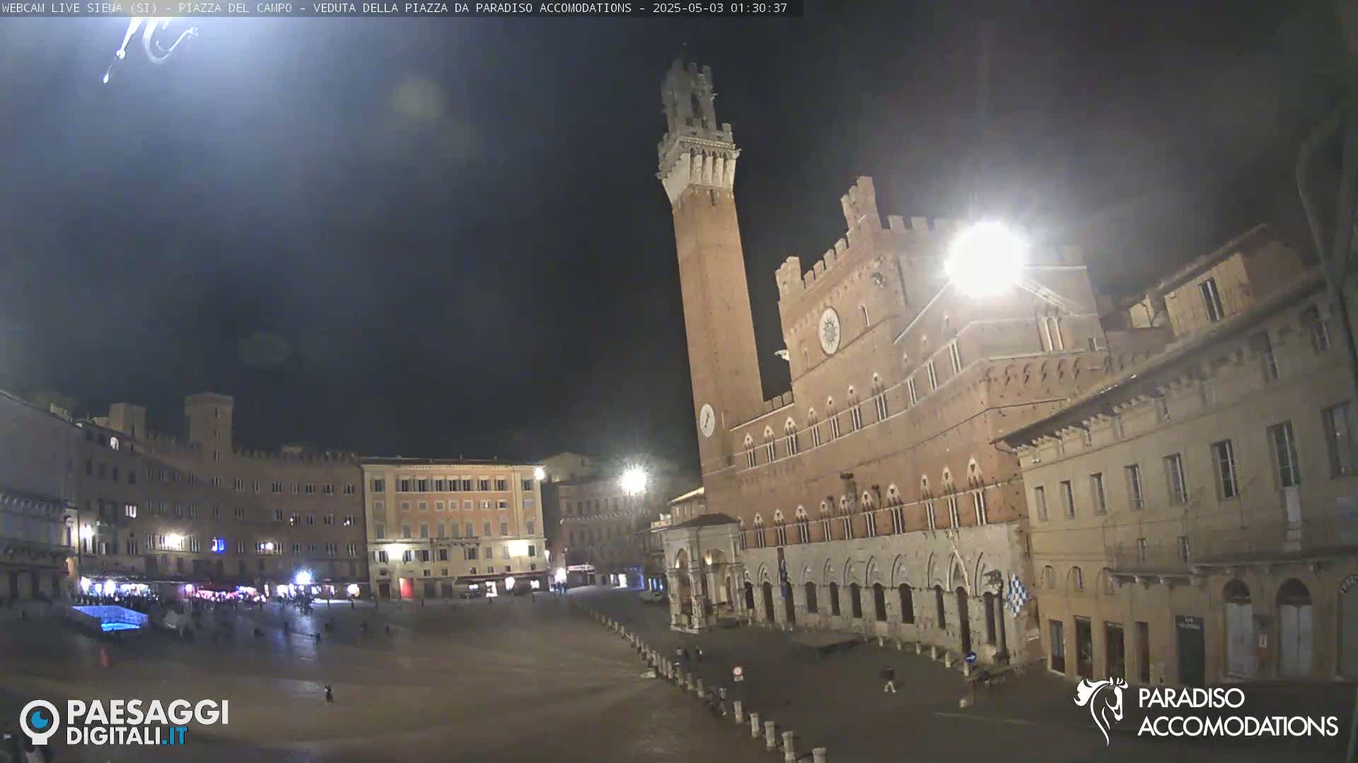 A nighttime view of a large, empty plaza in a European city, featuring a tall brick tower and surrounding buildings.