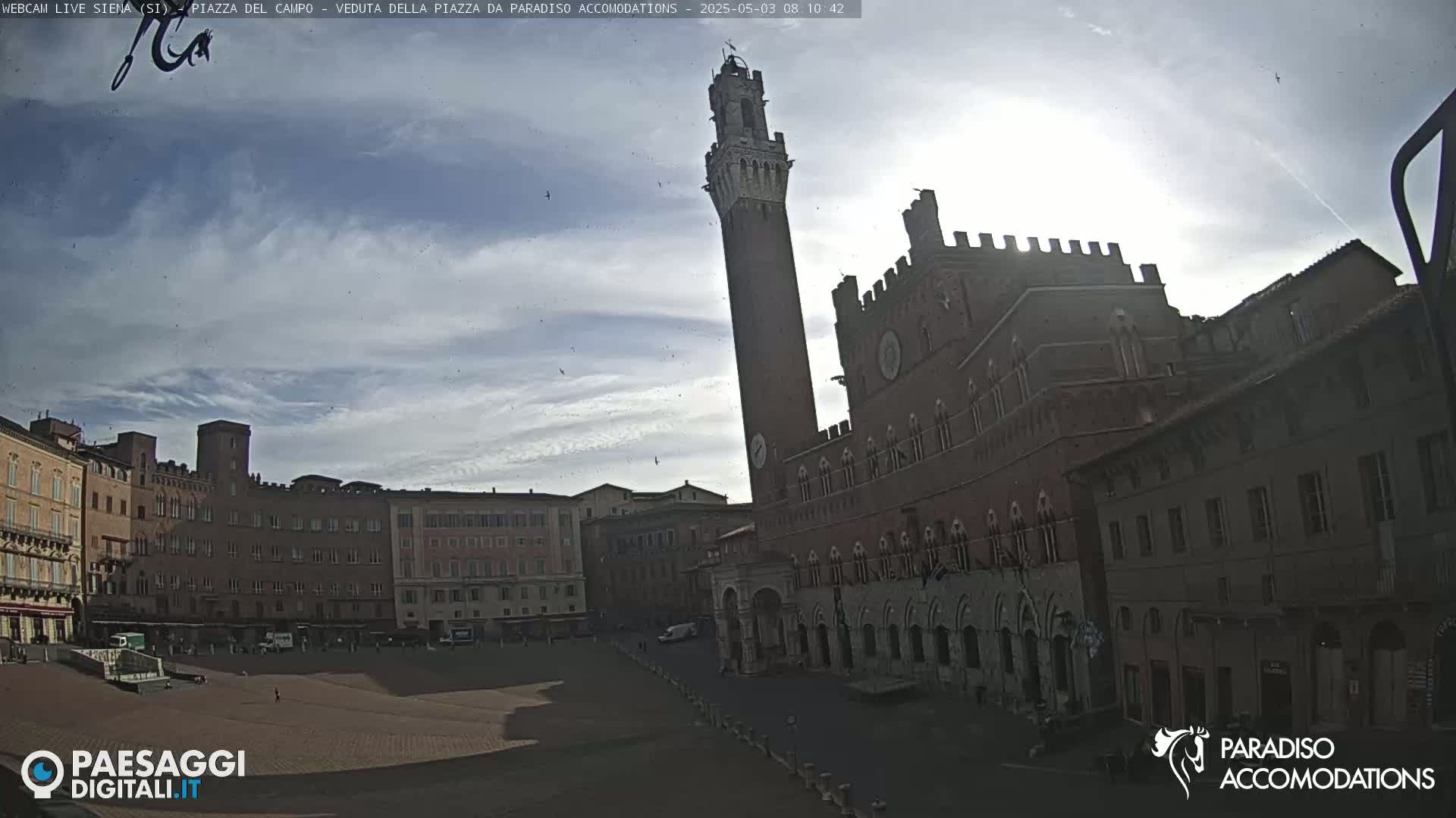 A mostly sunny day reveals a large, empty plaza surrounded by historic brick buildings, including a tall, slender tower.