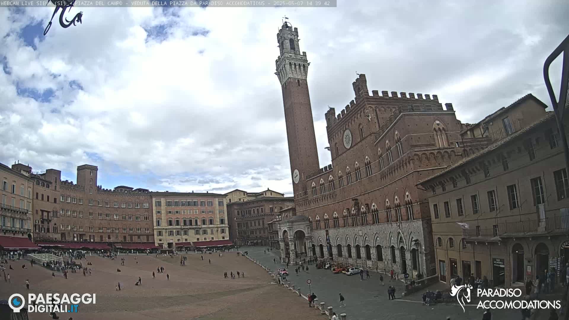 A large, mostly empty, brick plaza is surrounded by medieval-style buildings under a partly cloudy sky.