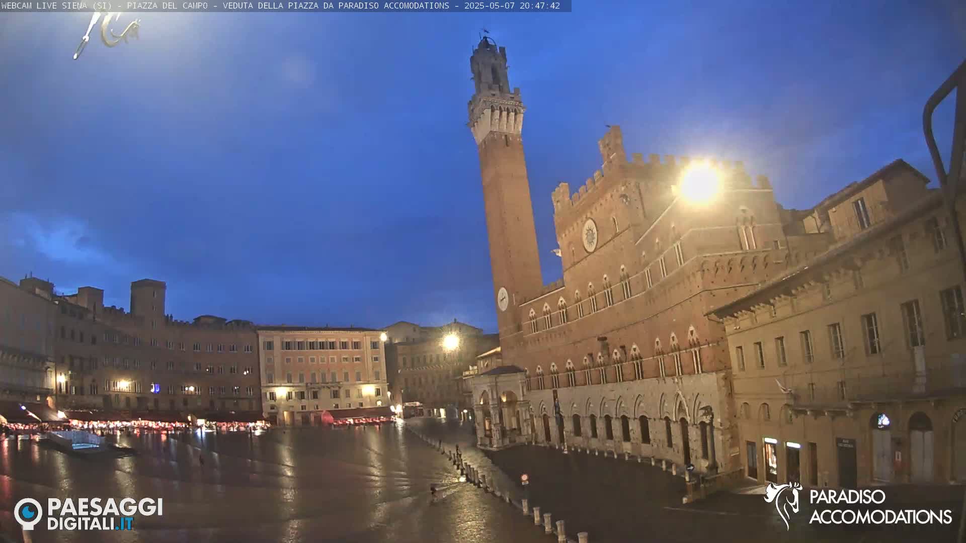 A large, mostly empty town square at twilight, featuring several brick buildings, one with a tall tower, under a clear, dark blue sky.