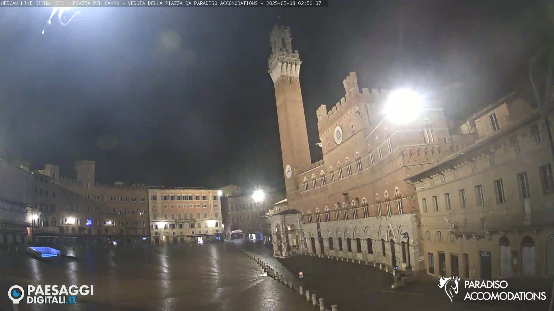 A large, empty, wet plaza at night is surrounded by historic brick buildings, including a tall tower.